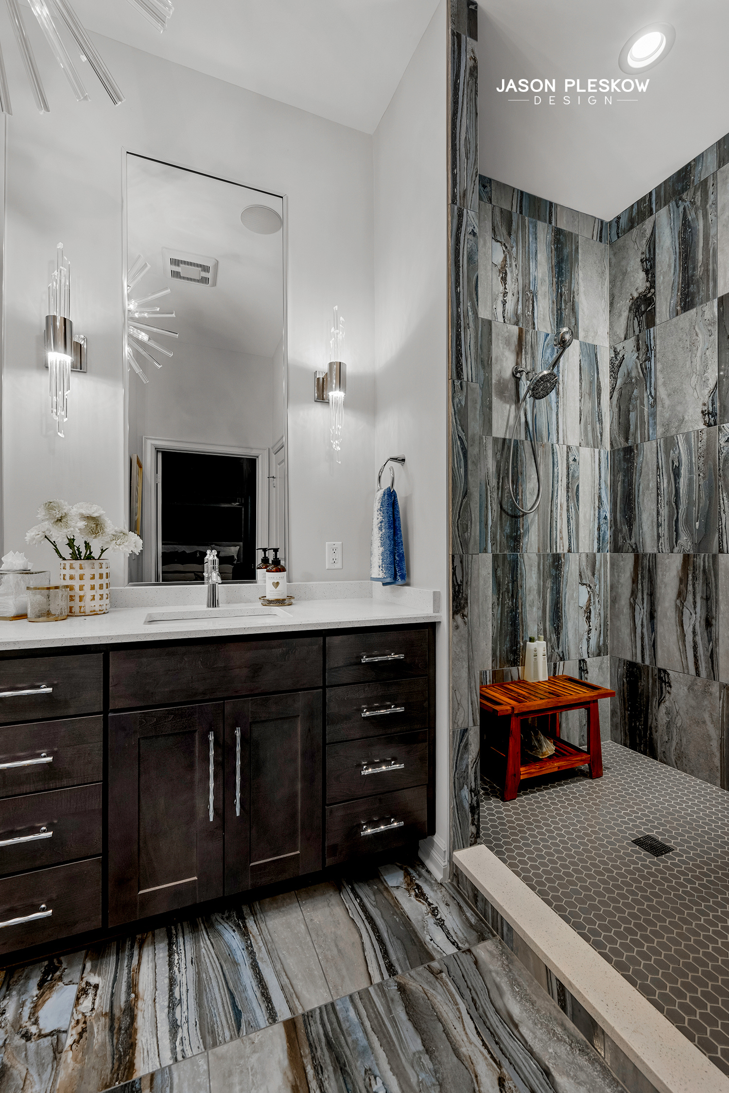 Modern bathroom with a dark wood vanity, white countertop, large mirror, decorative wall sconces, and a walk-in shower with gray and brown textured tiles and a small wooden stool.