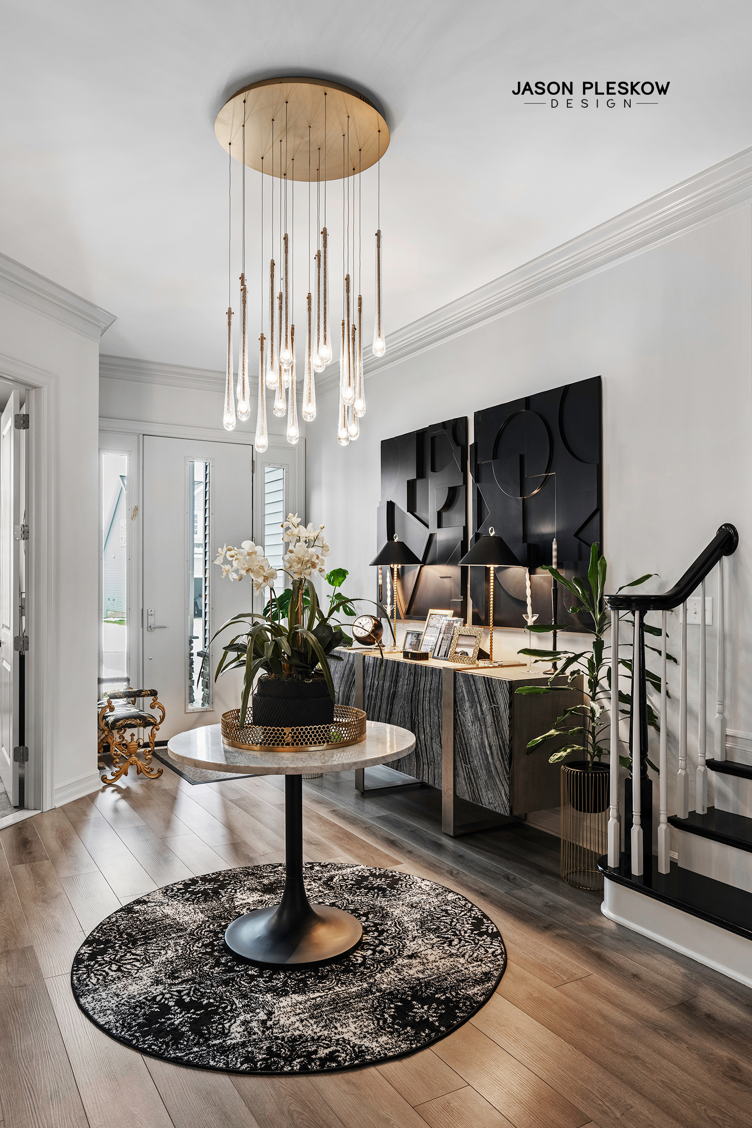 Modern entryway with a chandelier, black and gold accents, a black and white patterned rug, and a console with lamps and framed photos.