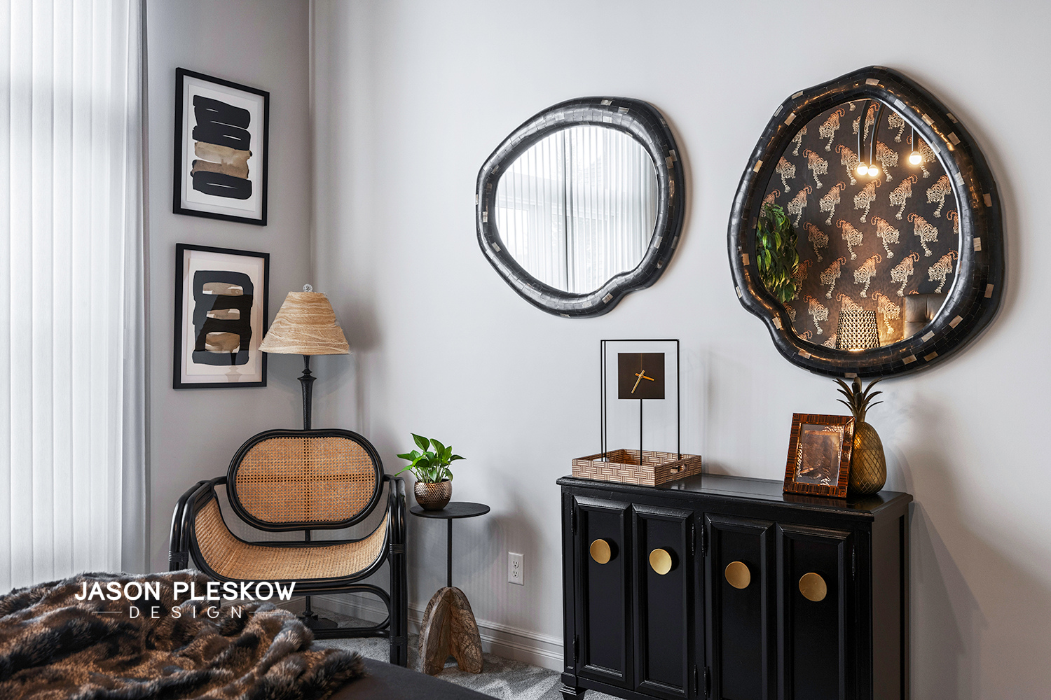 Living room corner with black cabinet, gold-accented pineapple mirror, two irregular black-framed mirrors, abstract artwork, rattan chair, lamp, small table with plant, and decorative items.