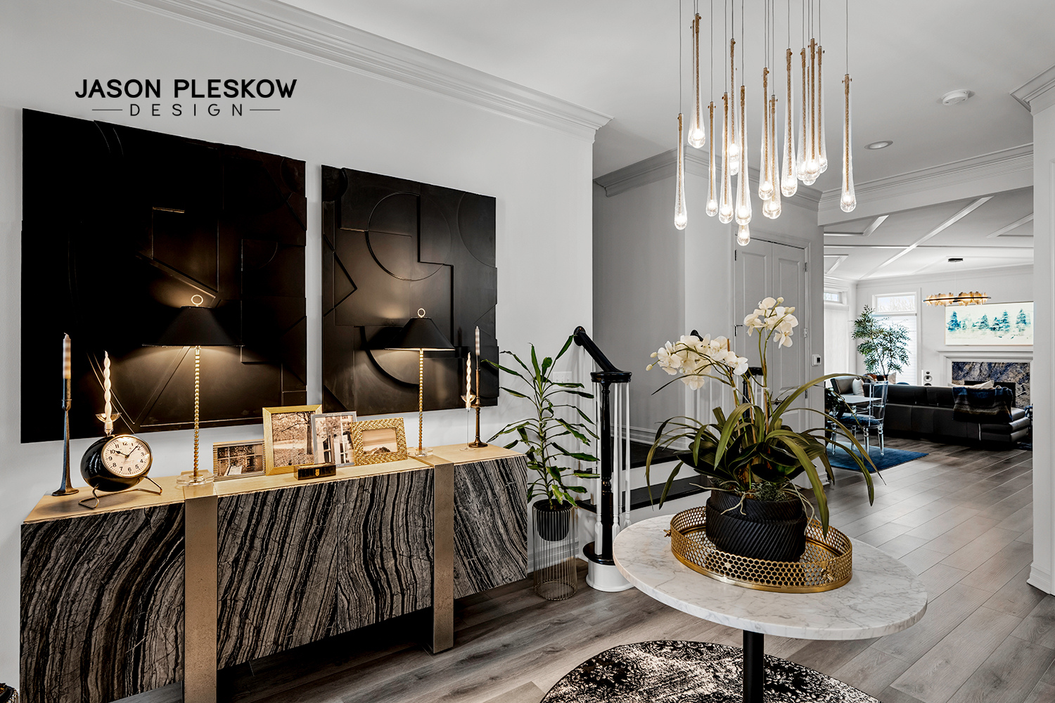 Modern living room with black and white decor, featuring a marble-top table with a potted orchid, a textured black cabinet with gold accents, and a hanging gold light fixture. There is a distant seating area with a fireplace and large windows.
