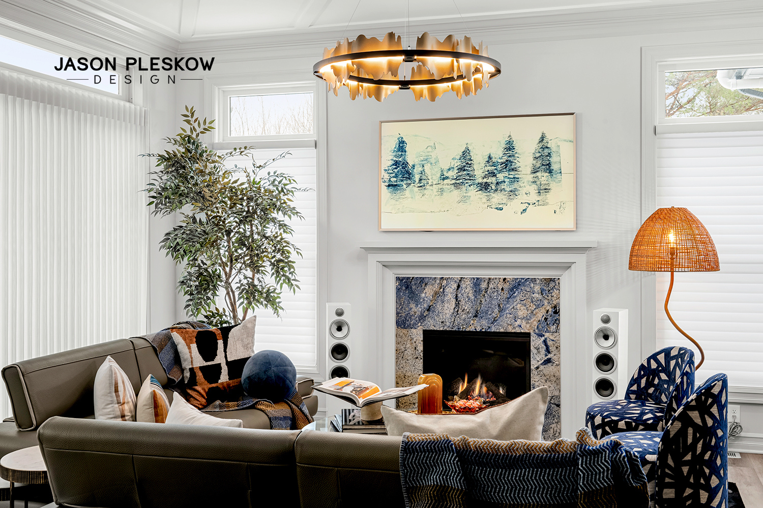 Modern living room with a fireplace, a large potted plant, a multicolored throw blanket, framed artwork above the fireplace, a rattan lamp, two blue patterned chairs, and a black leather sofa, with a circular chandelier overhead.