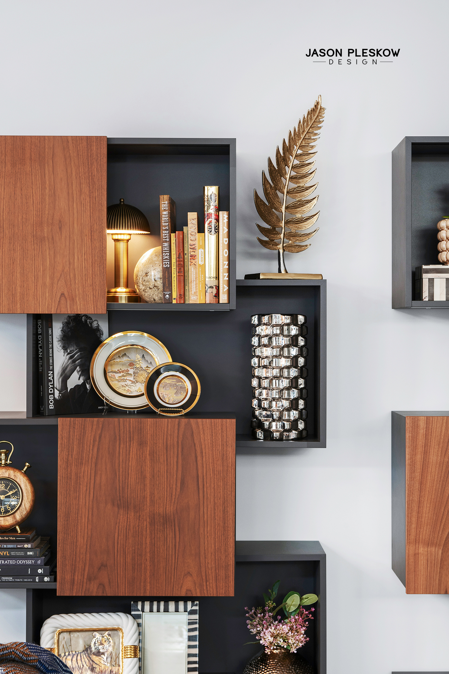 Decorative bookshelf with books, a gold lamp, metallic vase, framed plates, flowers, and a gold clock.