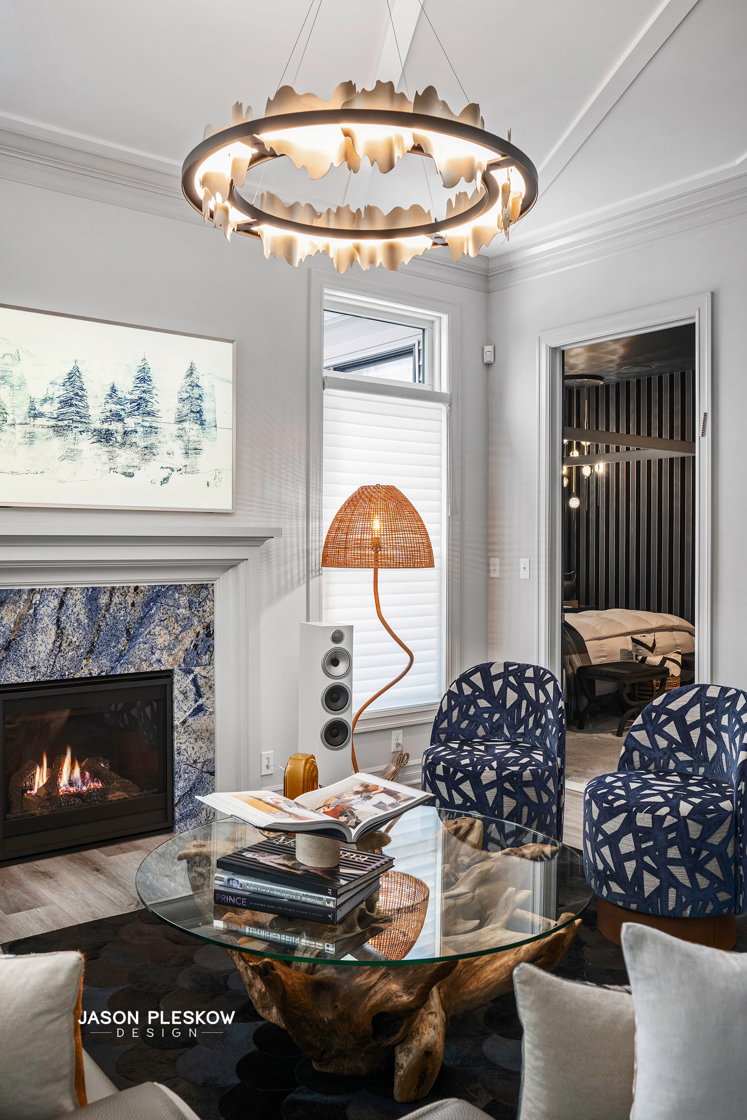Living room with a modern chandelier, fireplace with a stone surround, two blue patterned chairs, and a glass coffee table. A wicker floor lamp is beside a white window with blinds, and a partially visible doorway leads to a bedroom with a black stri