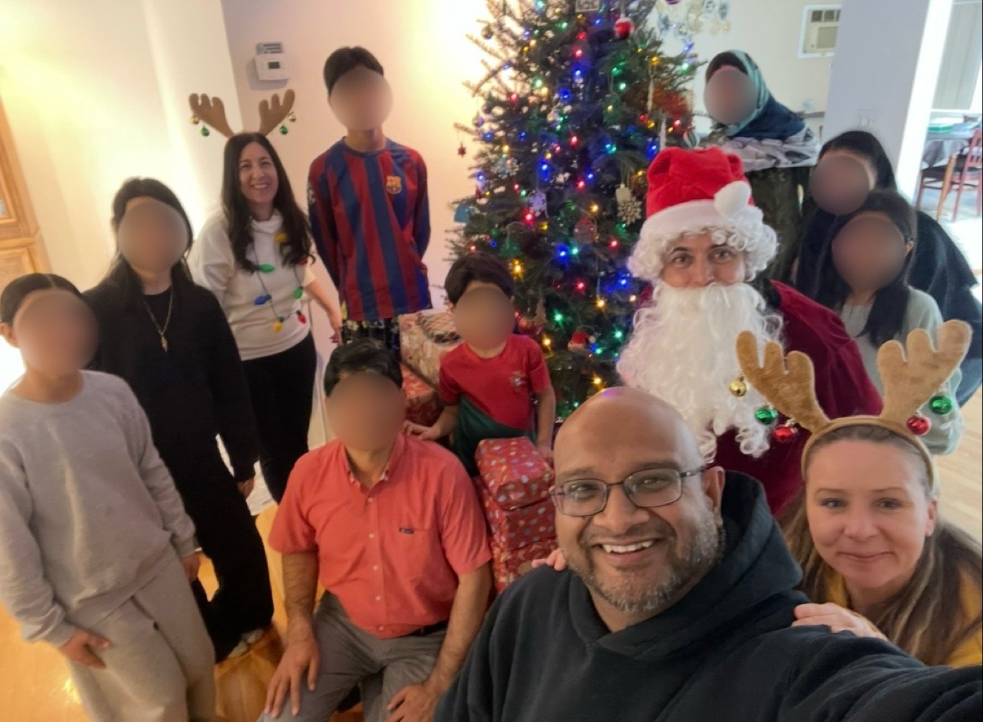 Family Christmas gathering with decorated wreath, Christmas tree with lights and ornaments, Christmas gifts, and people wearing holiday accessories including Santa hat, reindeer antlers, and Christmas sweater.