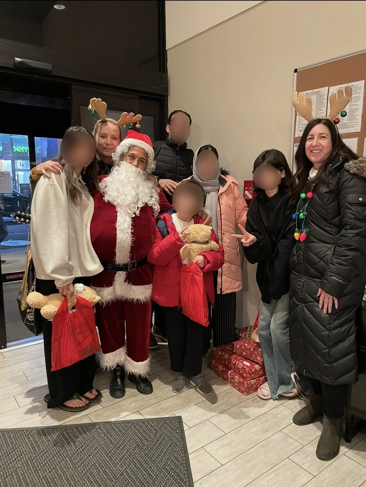Group of people, including children, gathered with Santa Claus dressed in a traditional red velvet suit with white trim, at a festive holiday event. Some individuals are wearing reindeer antler headbands. One child holds a teddy bear, and others hold