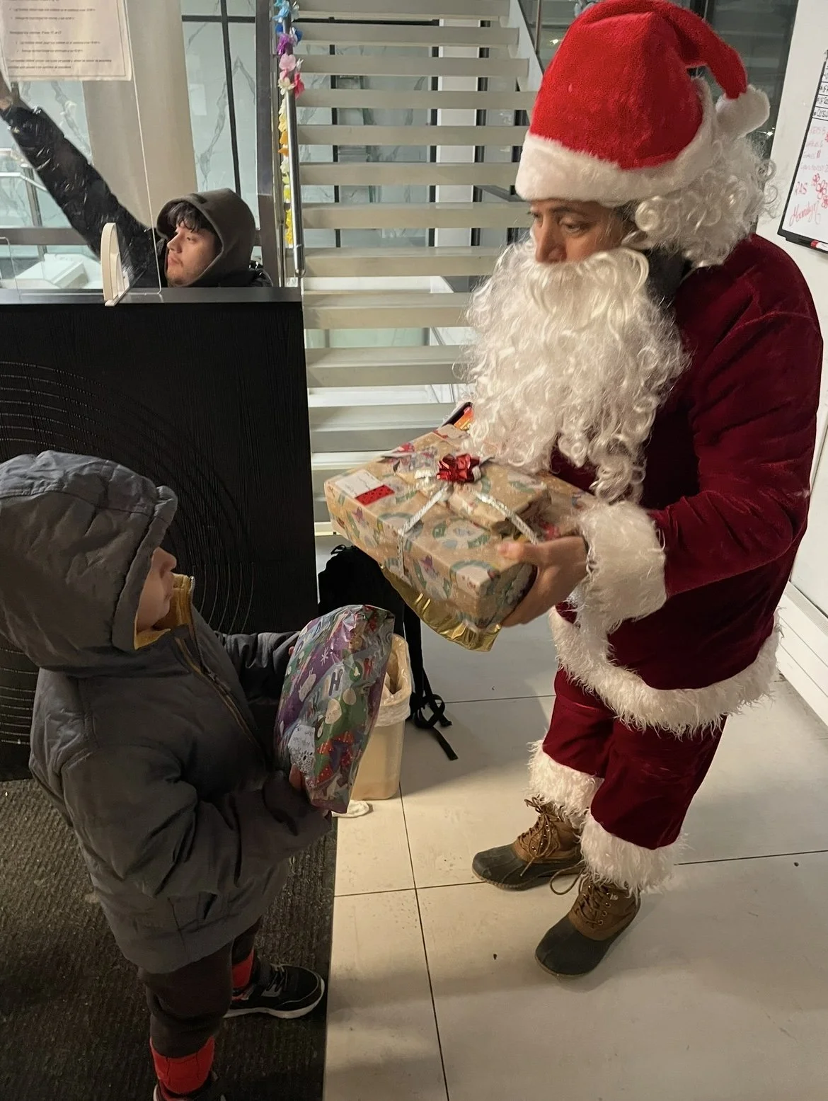 Santa Claus handing a wrapped Christmas gift to a young boy in a winter coat.  The boy holds a colorful gift bag and looks up at Santa.