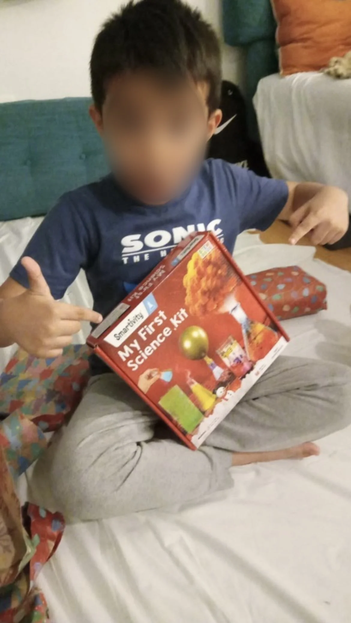 A young boy sitting cross-legged on a bed holding a science kit box labeled 'My First Science Kit,' pointing to the box, with colorful wrapped gifts nearby.
