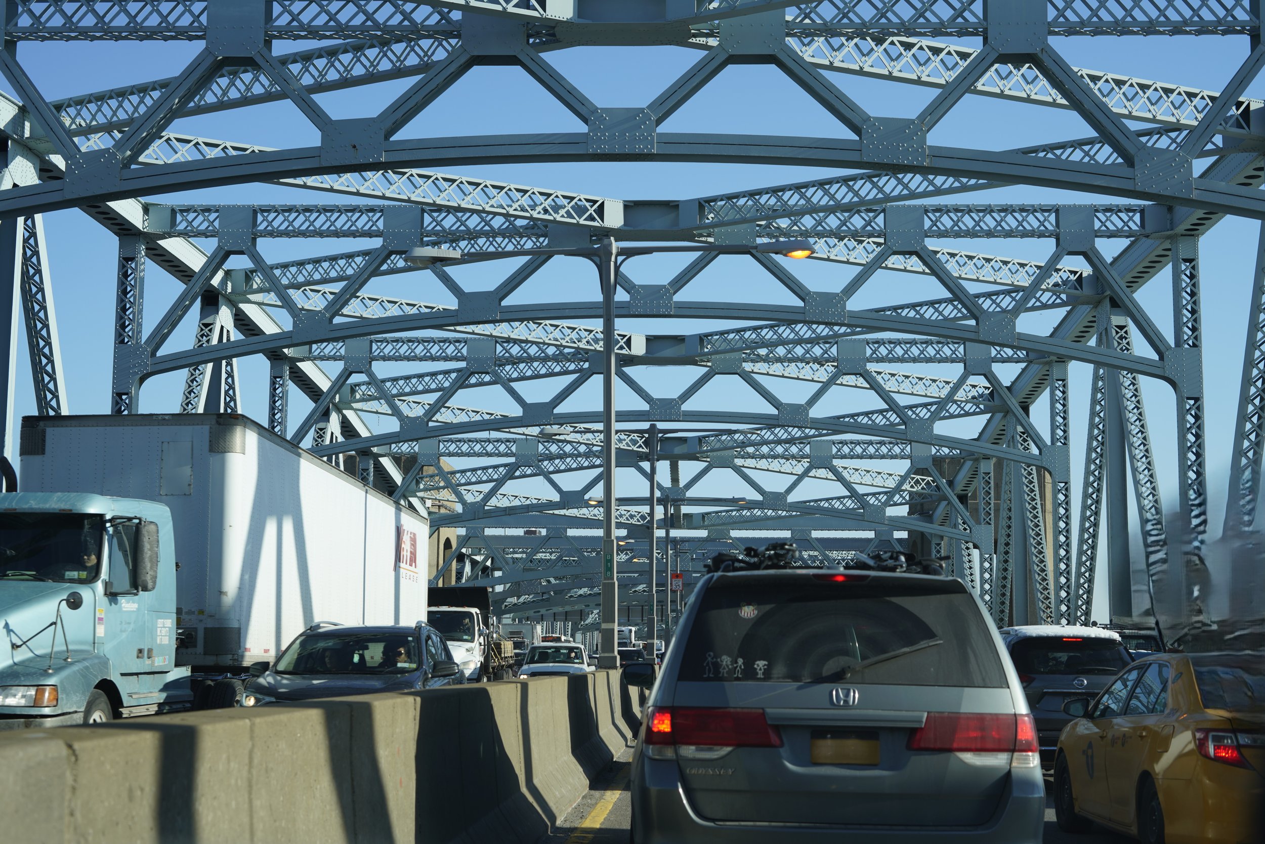 Line of vehicles on a bridge with a metal truss structure overhead, under a clear blue sky.