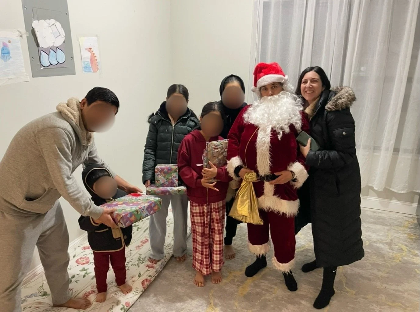 Group of six people, including a man dressed as Santa Claus, posing for a photo indoors during Christmas, with children holding gifts.