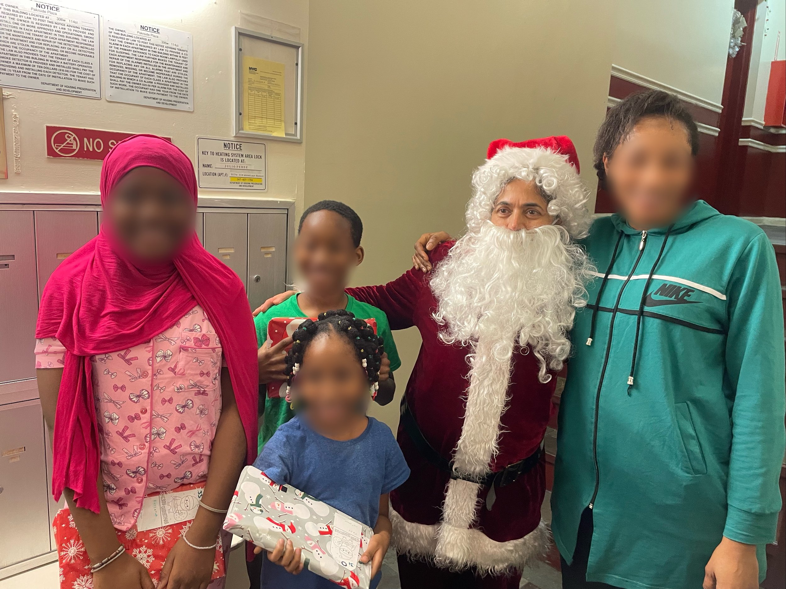 People celebrating Christmas in an indoor setting. Santa Claus is hugging a woman in a teal hoodie and a young boy holding a wrapped Christmas gift. Other children are also present, holding presents, with Christmas-themed wrapping paper. The woman an