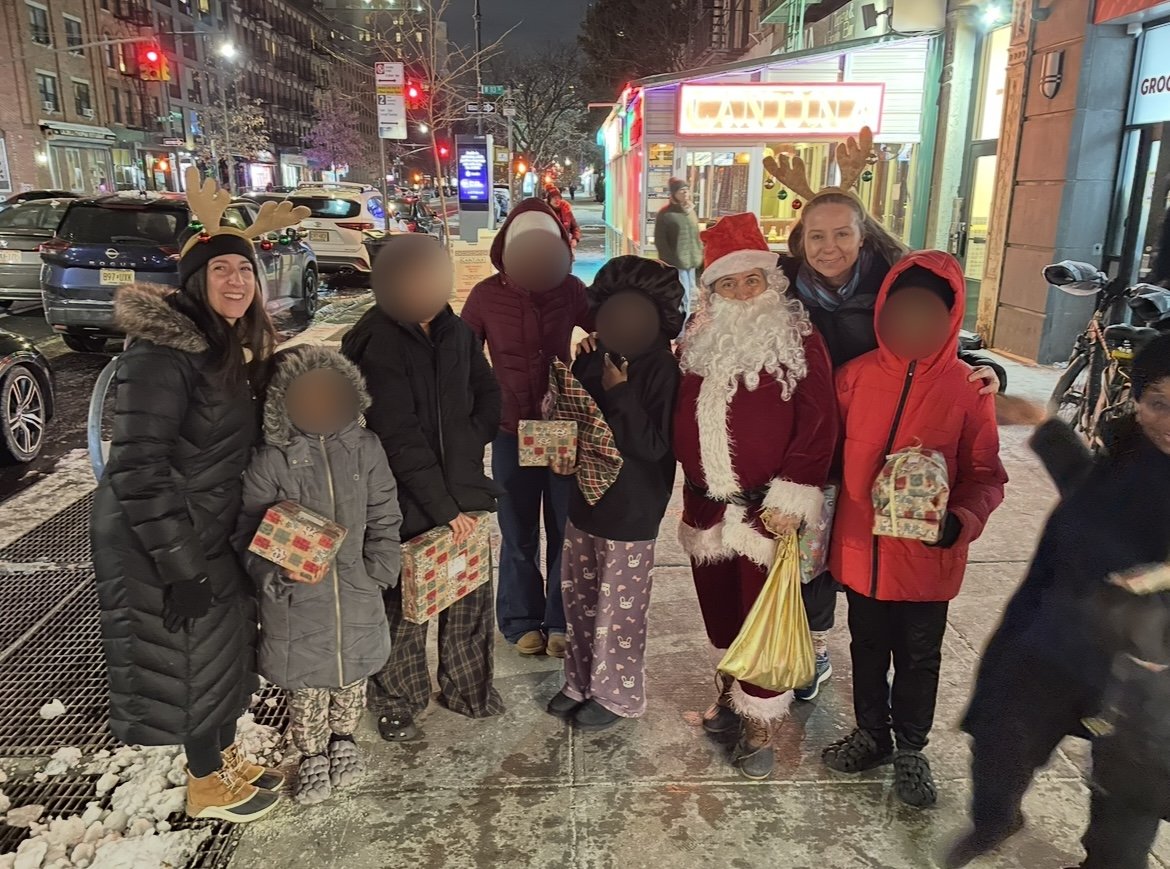 A group of people, including children and adults, standing on a city sidewalk at night. They are celebrating Christmas, with  Santa Claus and another woman wearing reindeer antlers. Some children are holding wrapped presents. In the background, there