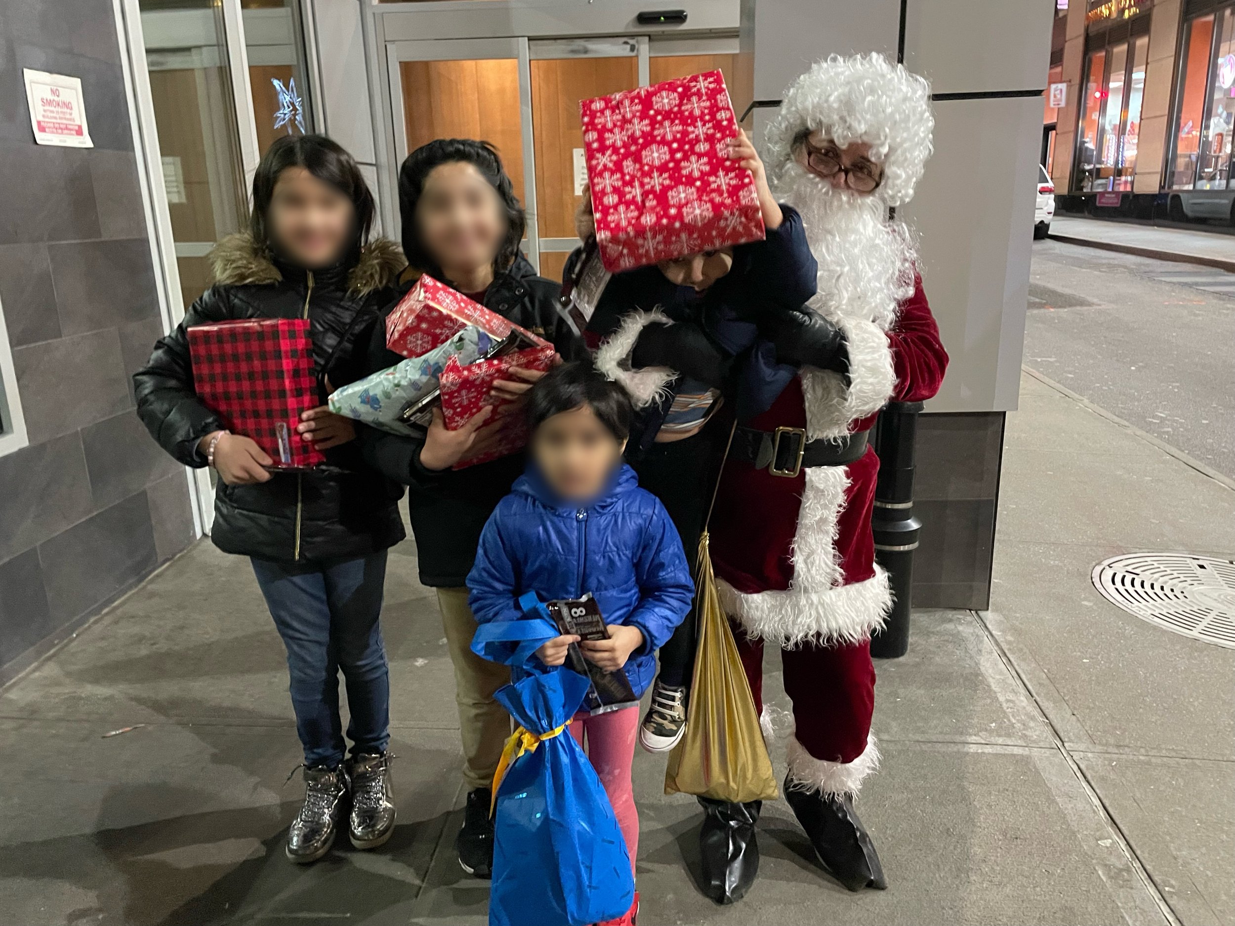 A group of children with blurred faces, standing near Santa Claus holding a large red gift bag, outside a building at night.