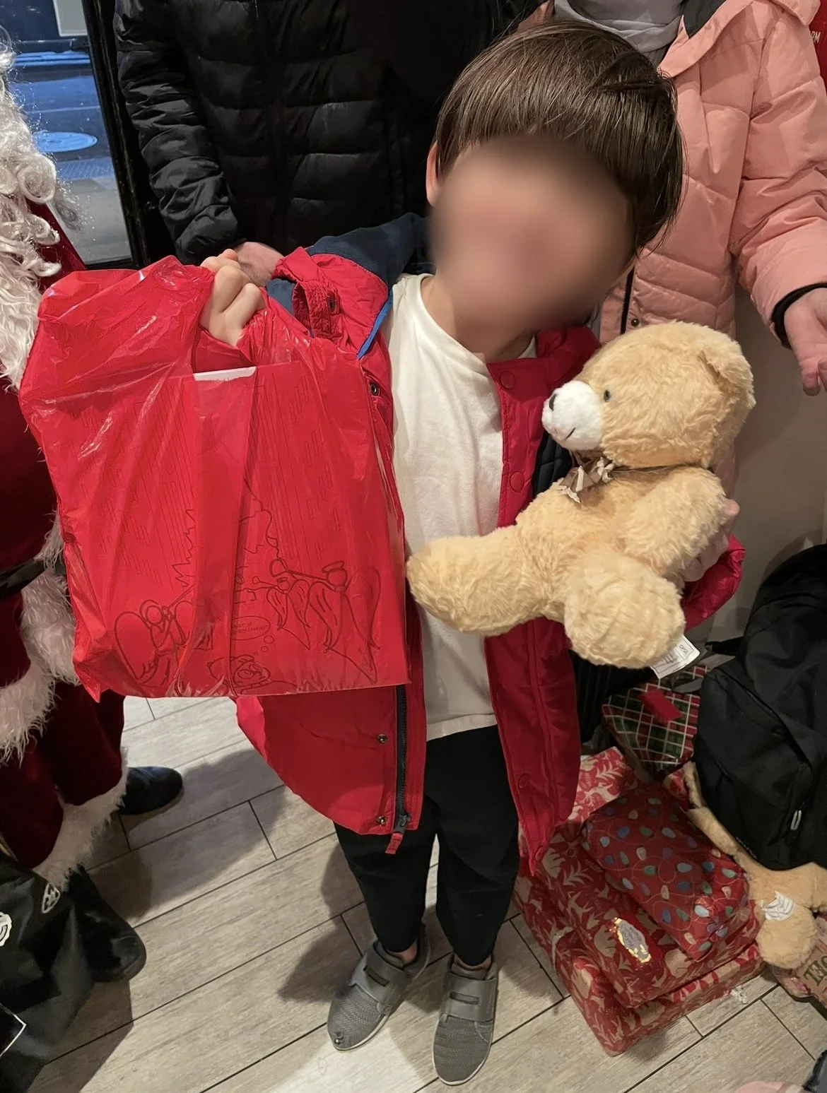 A boy holding a teddy bear with both hands, wearing a red jacket, surrounded by people, some in Christmas or winter attire, in a store decorated for Christmas with wrapped presents.