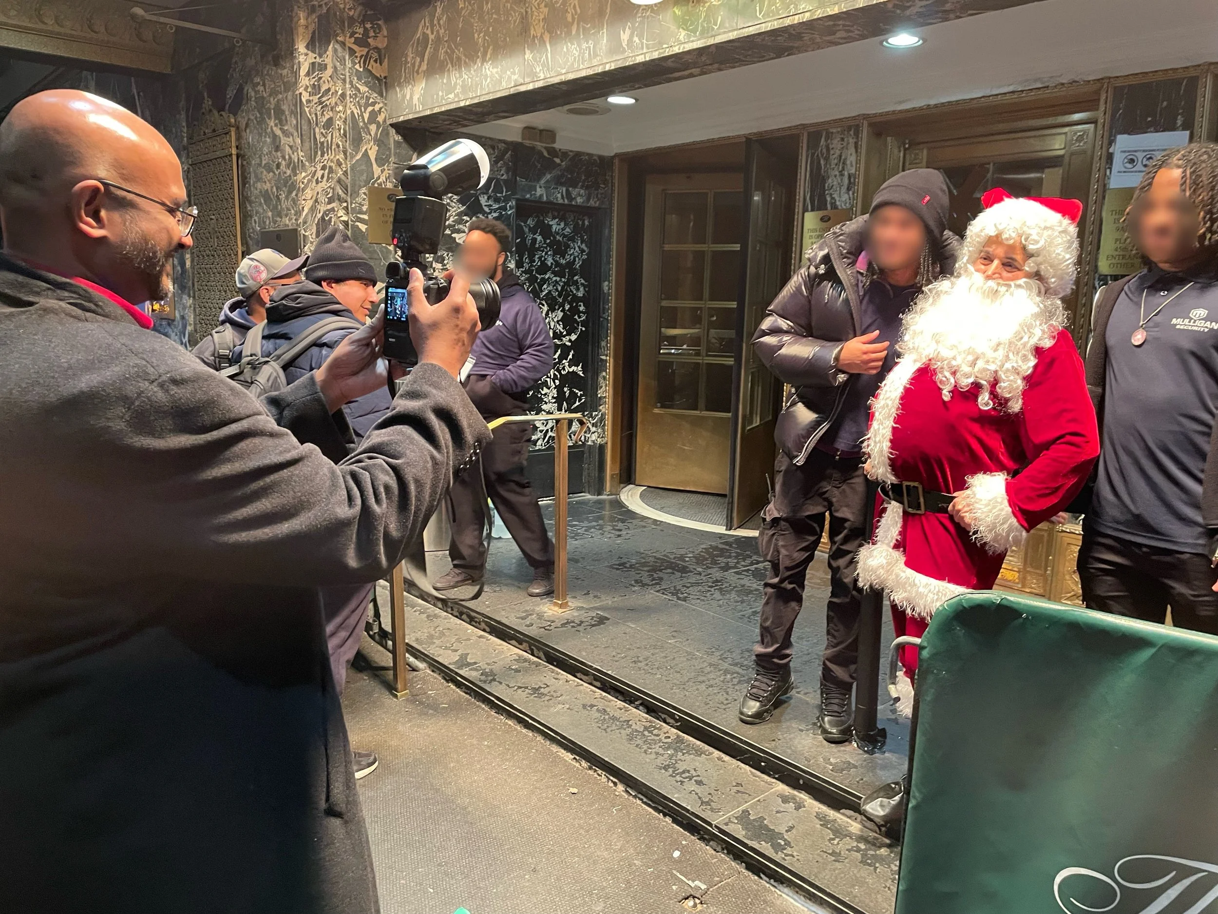 A group of people, some wearing winter clothing, posing with Santa Claus in a lobby or entrance of a building with dark marble walls. One person is taking a picture of the group.