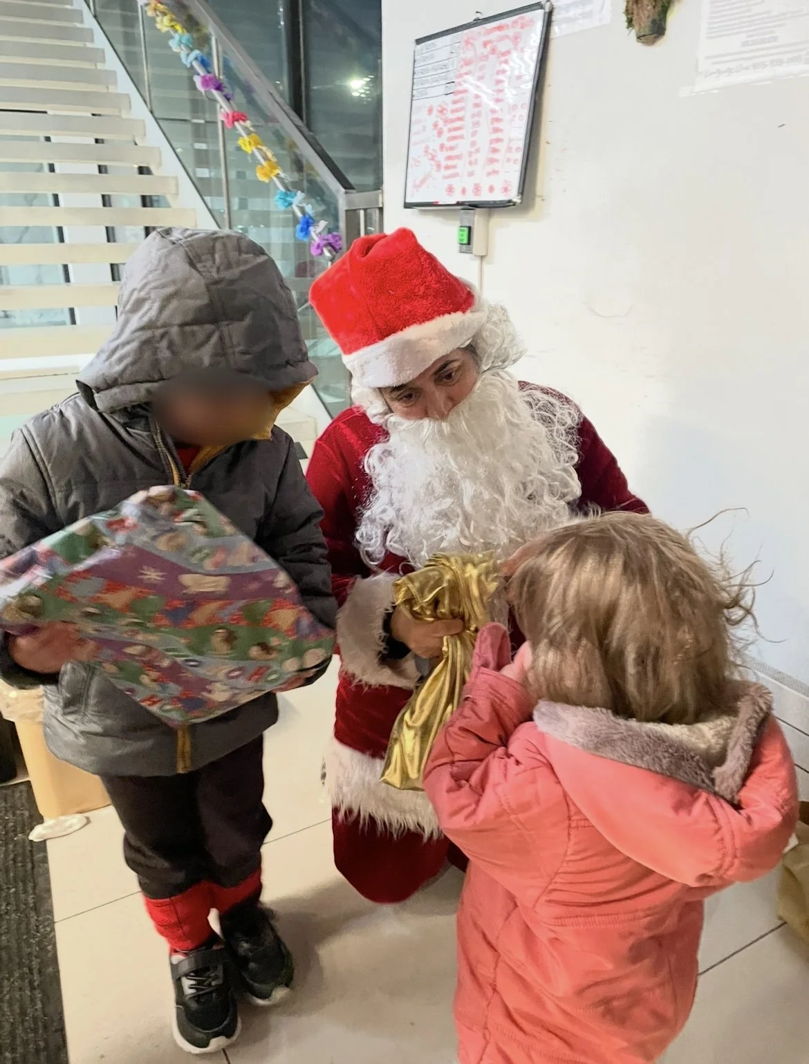 Santa Claus giving gifts to children indoors, with one child holding a present and another child talking to Santa.