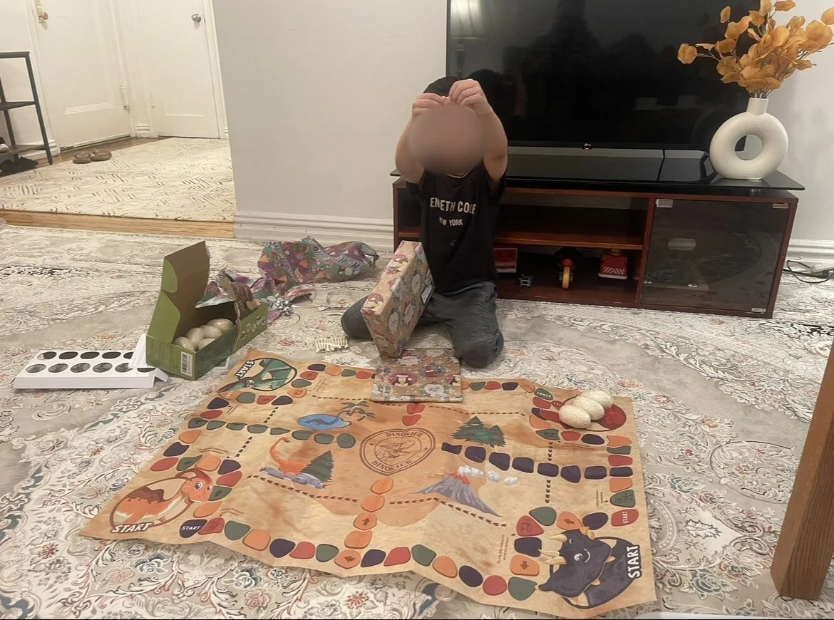 A child is playing a board game on a patterned rug in a living room. The child is kneeling and holding a box, with other game pieces and objects scattered on the rug.