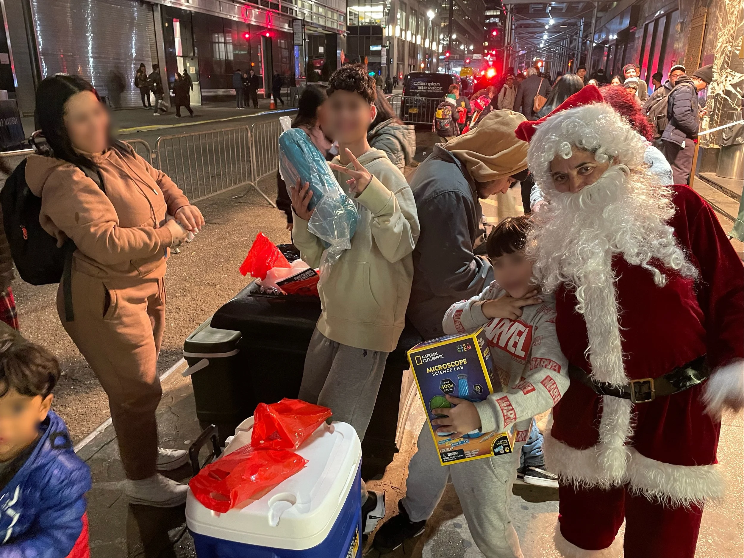 People gathered on a city sidewalk at night, some wearing coats and winter hats, others in festive attire including Santa Claus in a red velvet suit with a white beard, and children holding gift boxes. The scene is illuminated by streetlights and bui