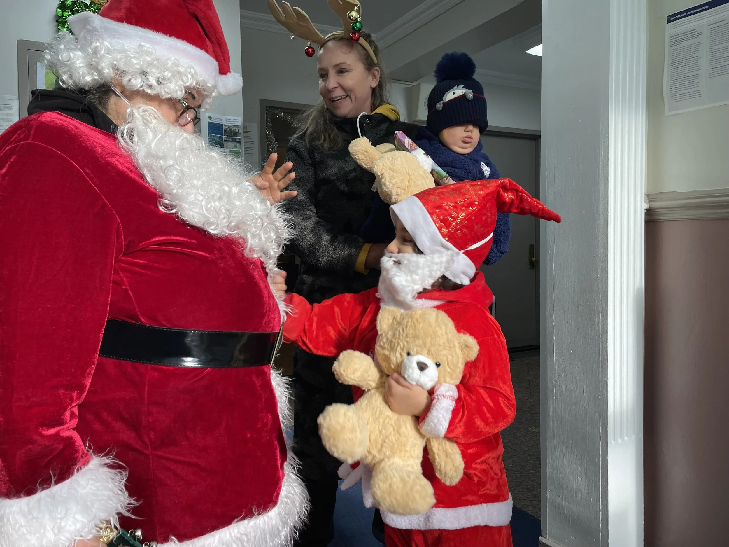  Santa Claus talking to a woman with reindeer antlers and two children in holiday outfits, one holding a teddy bear, indoors with holiday decorations.