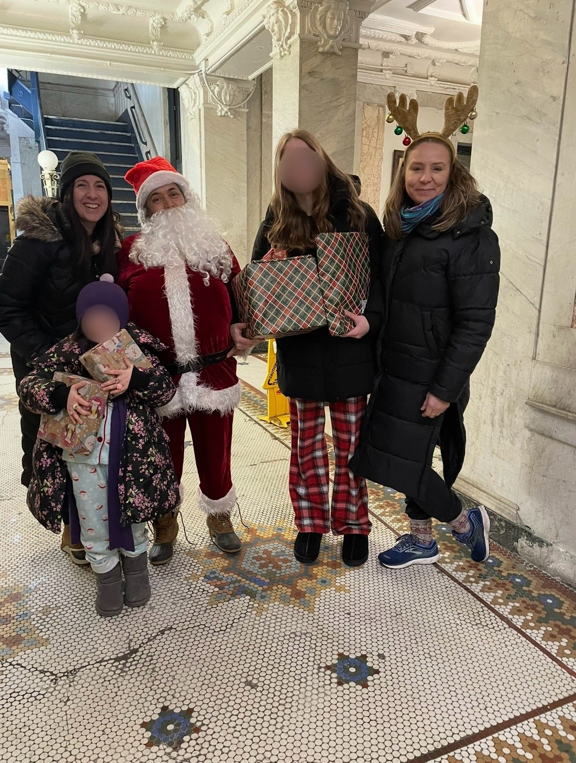 A group of people in holiday attire, including a person dressed as Santa Claus, standing in a festive indoor setting. They are holding wrapped Christmas presents, and one person is wearing reindeer antlers with ornaments. The group includes children 