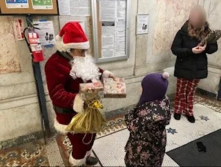 Santa Claus handing a wrapped gift to a child in a purple hat in a hallway or lobby.
