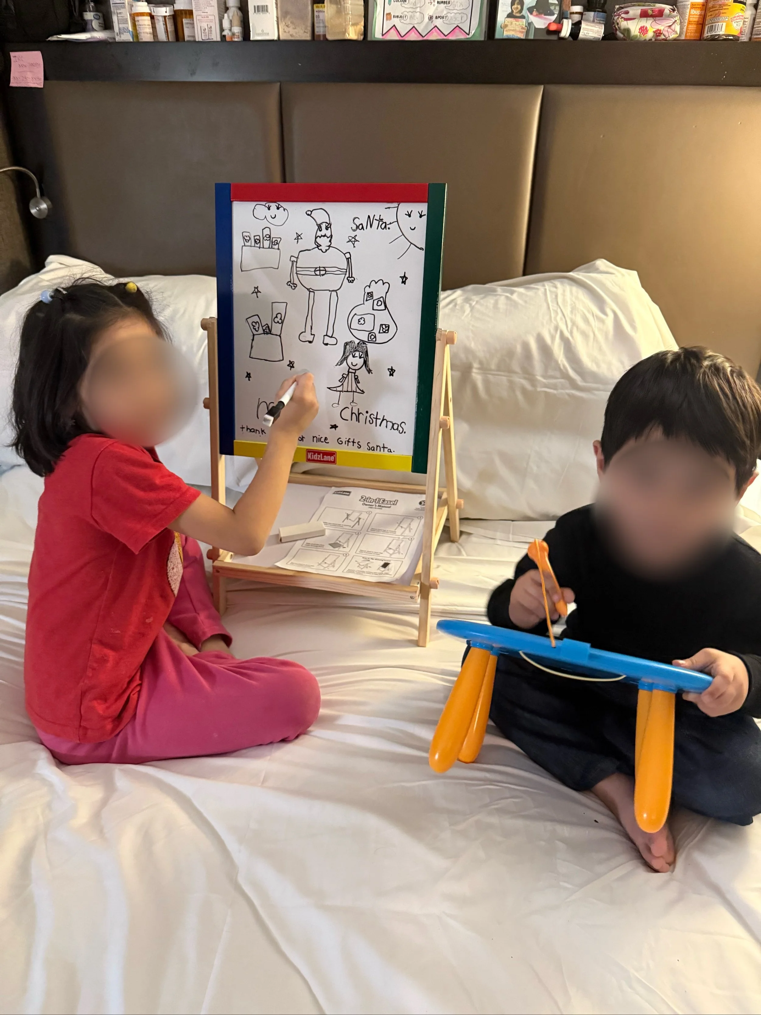 Two children sitting on a bed, one girl in a pink outfit drawing on a whiteboard and a boy in black with orange shorts playing with a magnetic drawing board. The whiteboard has a Christmas drawing with Santa, a girl, and various Christmas-themed item