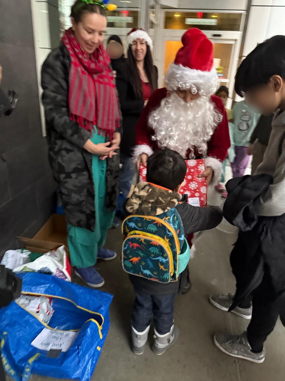 Children and adults gathered around a person dressed as Santa Claus handing out gifts, with Christmas hats and festive clothing.