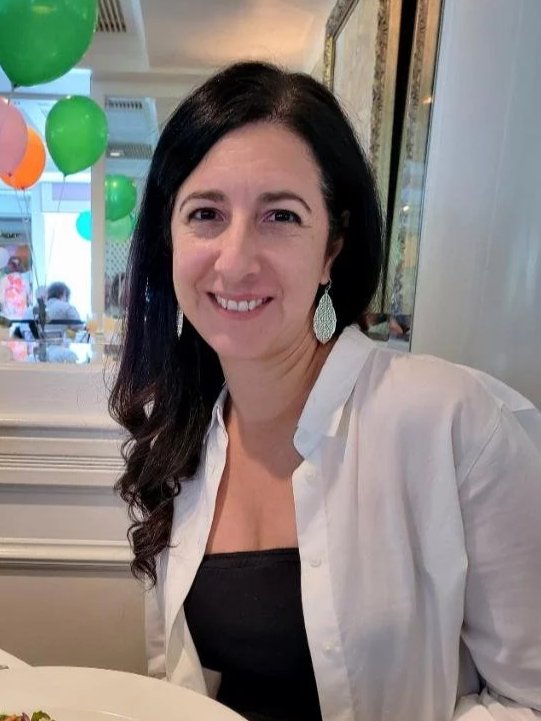 A woman with long dark hair wearing a white shirt and teardrop earrings, smiling at a celebration with green and orange balloons in the background.