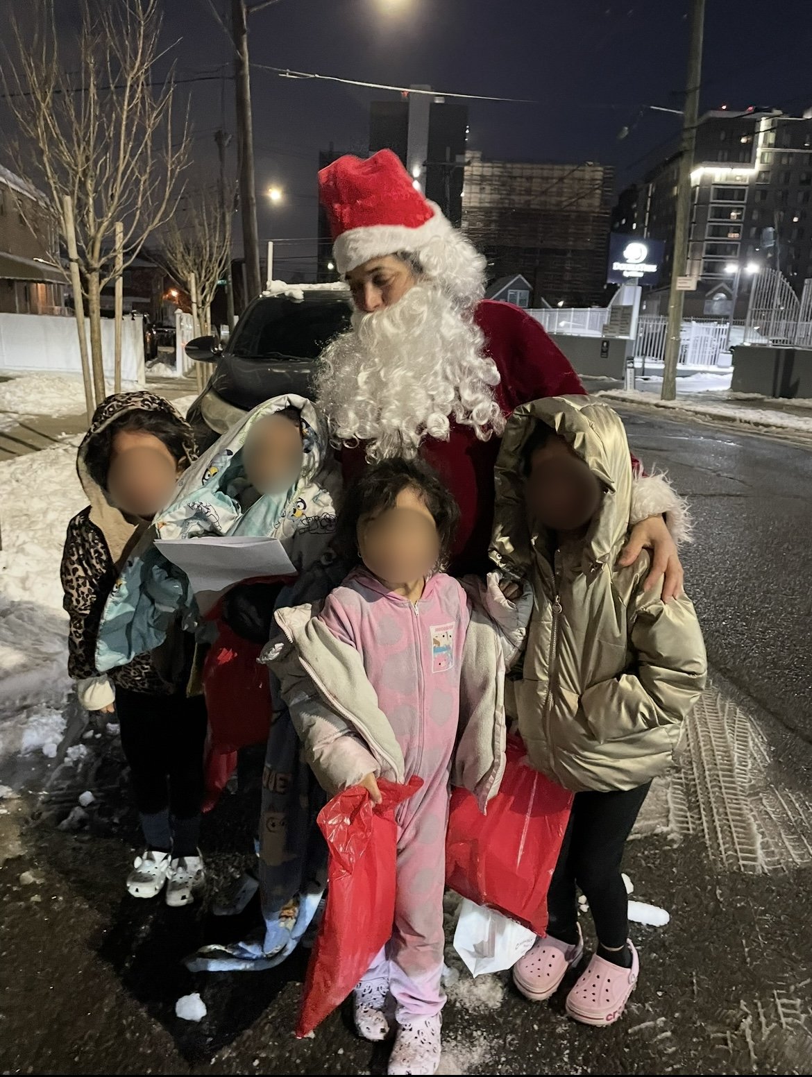 Santa Claus surrounded by four children in winter clothing, standing on a snowy street at night.
