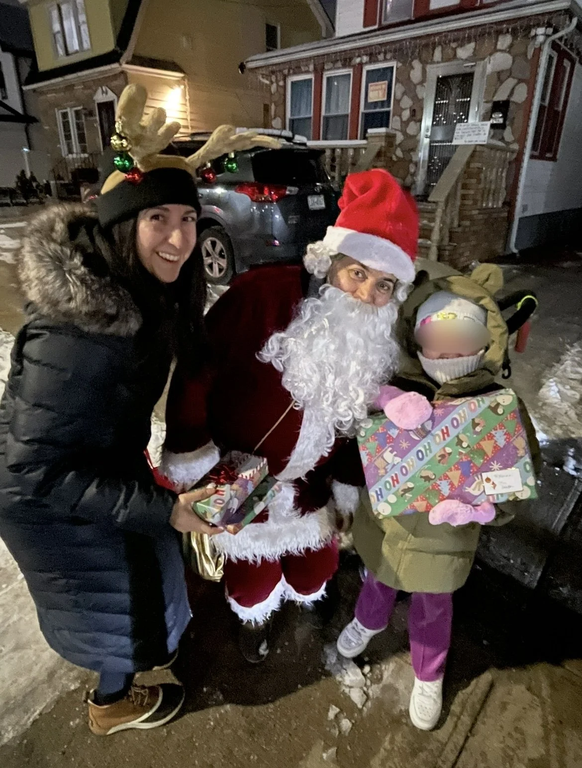 A woman and a girl standing with Santa Claus outside at night. The woman is wearing a black coat with a fur-lined hood and a Christmas hat with reindeer antlers. The girl is holding a wrapped gift and has her face blurred, wearing a green coat, pink 