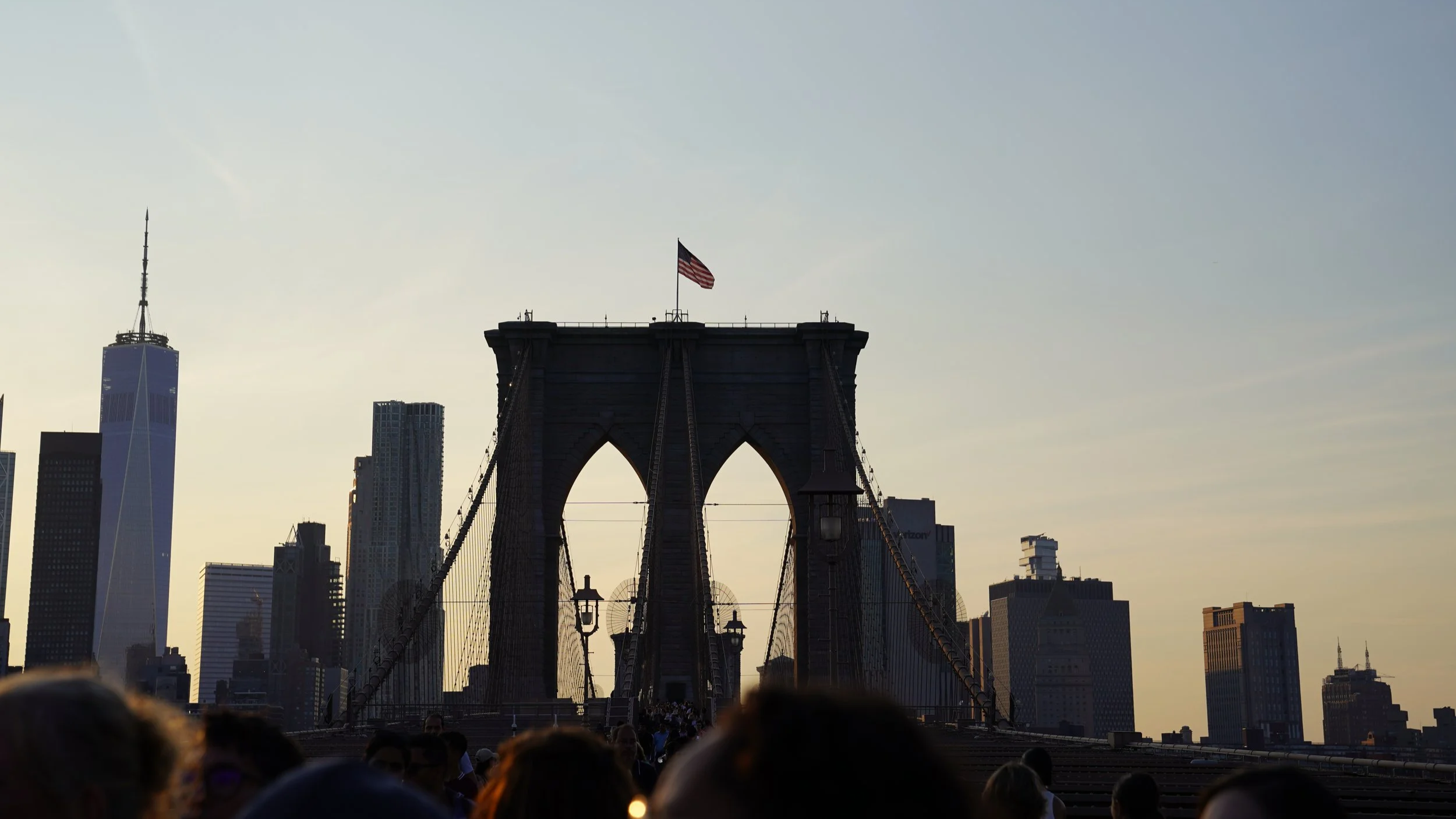View of the Brooklyn Bridge with the New York City skyline, including One World Trade Center, in the background at sunset.
