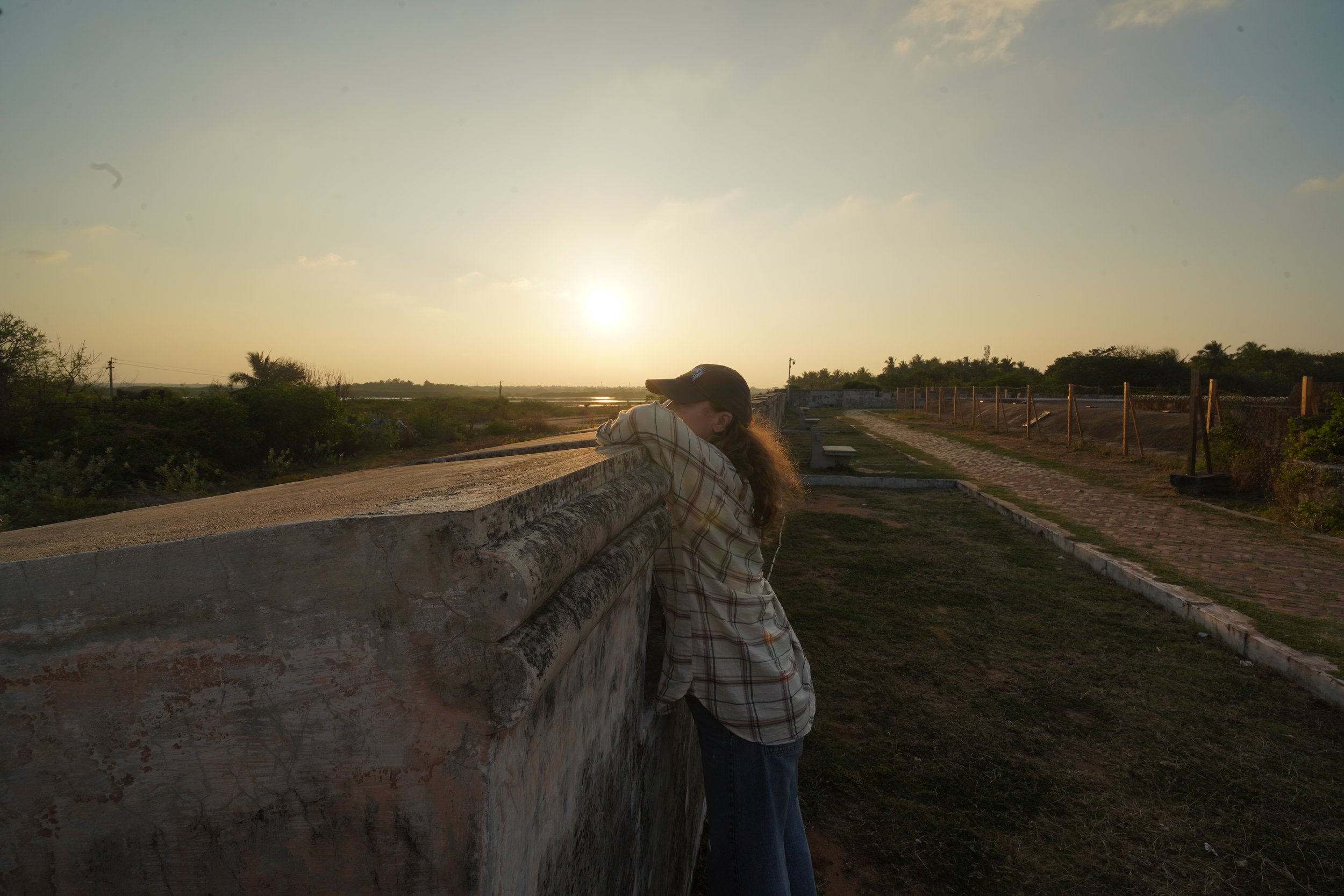A woman leaning on a stone wall during sunset, wearing a plaid shirt and a cap, with greenery and a pathway in the background.