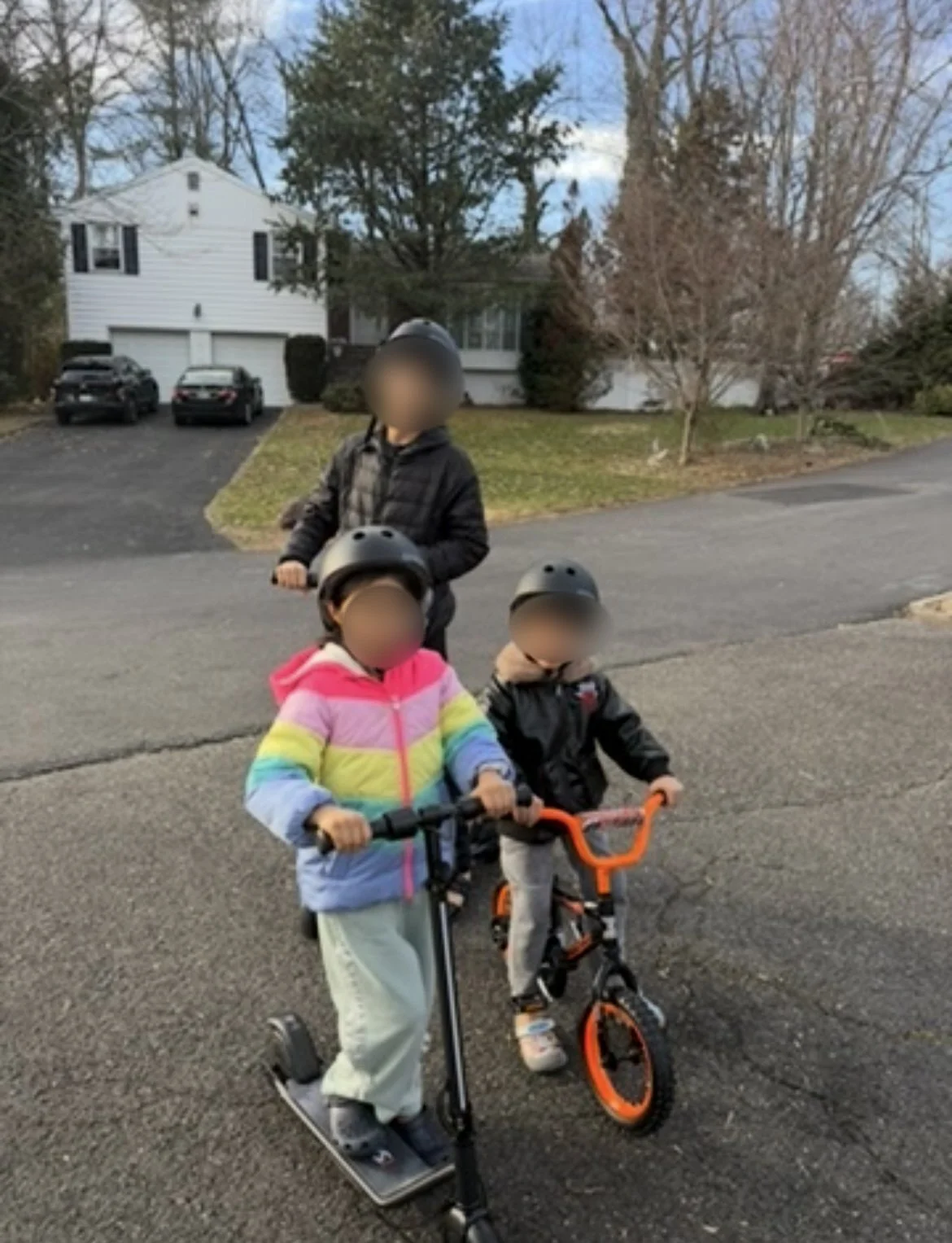 Three children wearing helmets and riding scooters and a bike on a residential street during daylight, with a white house and parked cars in the background.