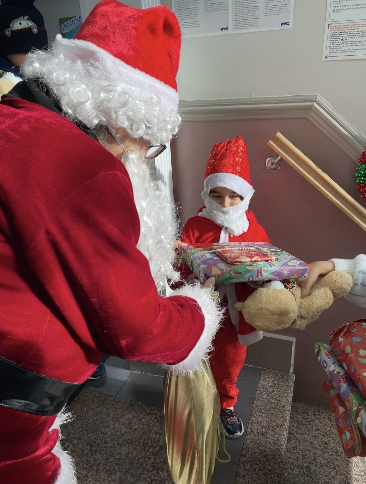 A child dressed as Santa Claus receiving a wrapped Christmas gift from Santa Claus. The child is holding a teddy bear and smiling at the gift exchange.