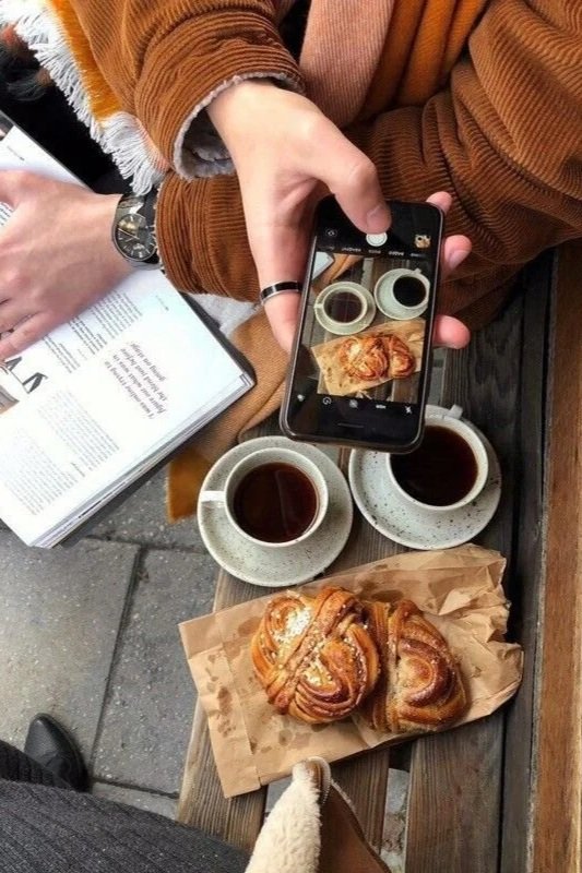Person taking a photo of a pastry with a smartphone. On the table, there are two cups of coffee and three pastries on a paper napkin. The person is wearing a brown jacket and has a ring on their finger.
