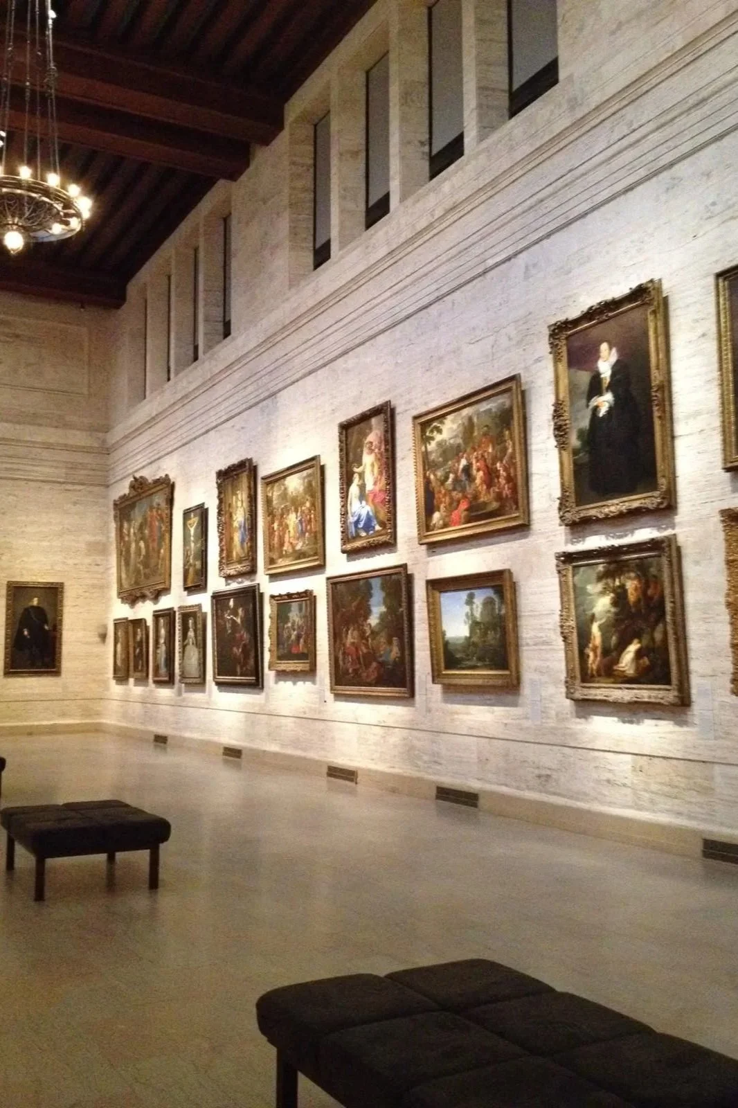 Interior view of an art gallery with a white brick wall displaying numerous framed classical paintings, some depicting people and outdoor scenes, with benches in the foreground.