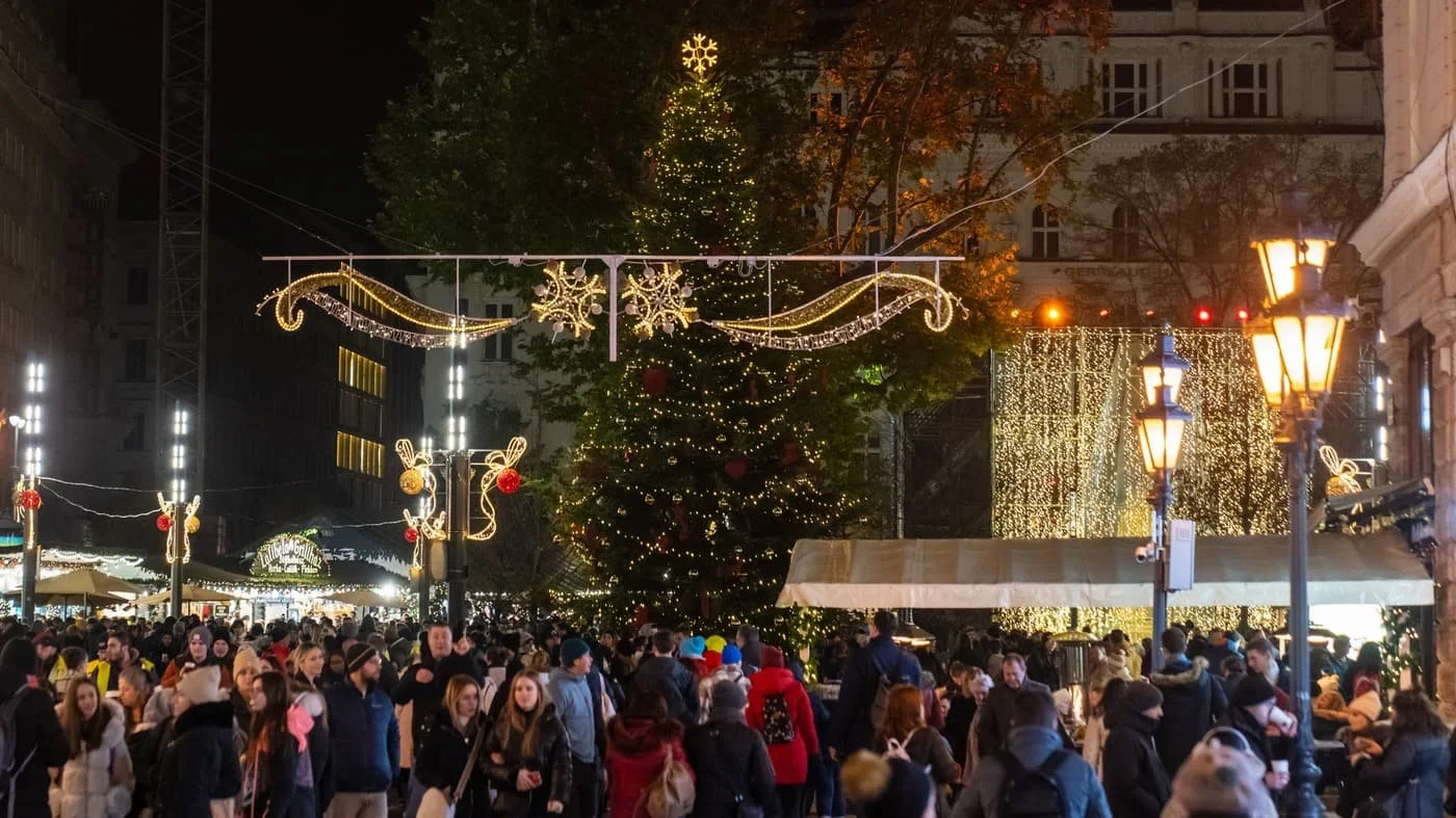 A crowded Christmas market at night with a large decorated Christmas tree in the center, surrounded by festive light displays and string lights, with many people walking and enjoying the holiday atmosphere.
