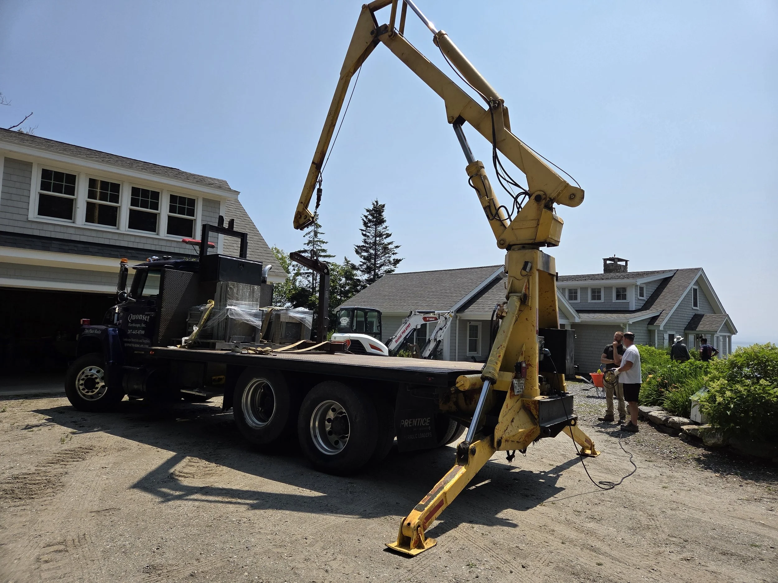 A construction truck with a yellow crane arm parked on a driveway in front of houses. Two men are talking near the house on the right, with construction equipment nearby.