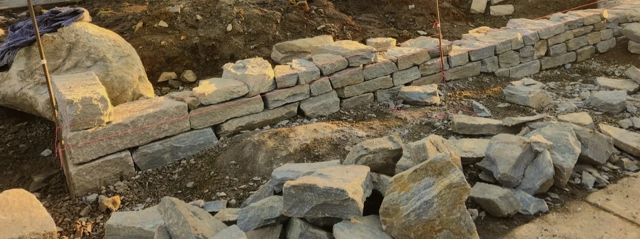 A partially built stone wall on a construction site with stones and dirt around, with a measuring string stretched along the wall.