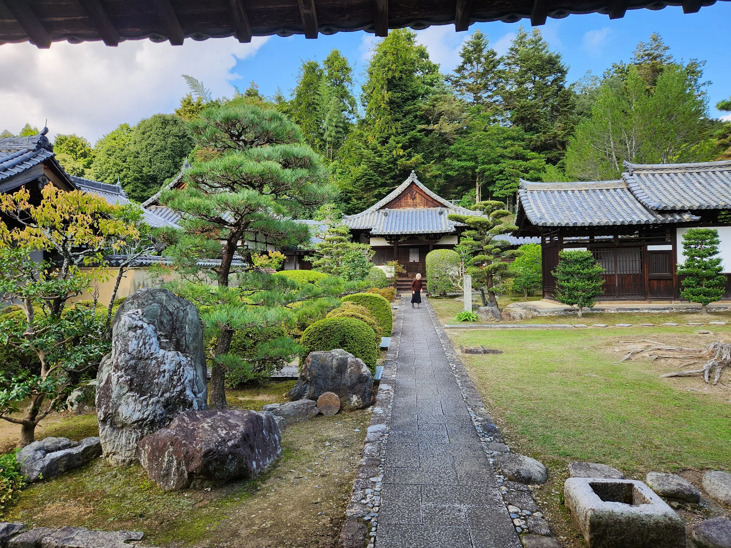 Traditional Japanese temple courtyard with stone pathway, manicured trees, rocks, and wooden buildings, surrounded by lush greenery and a blue sky.