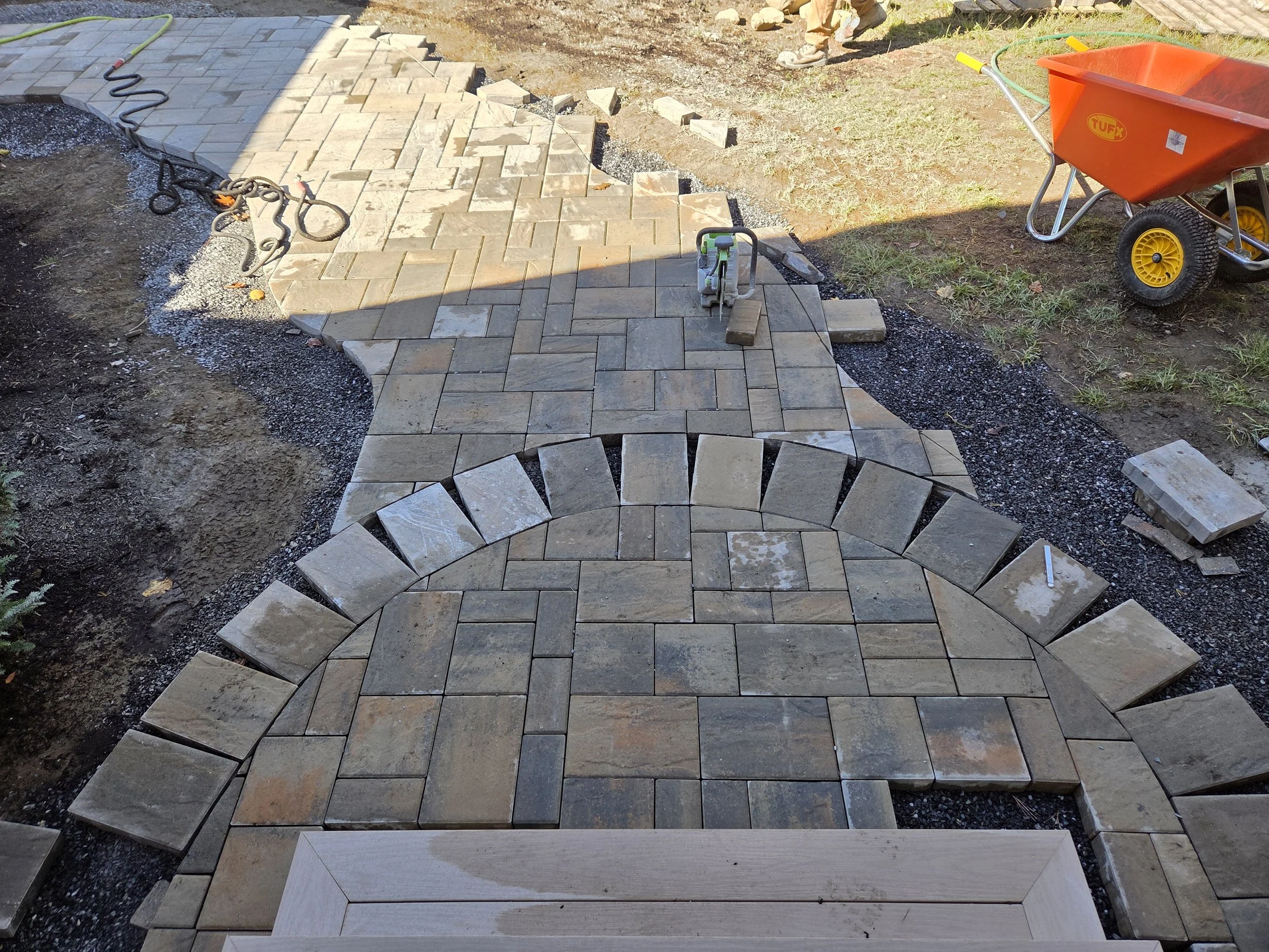 Paving stones being installed in a pattern for a walkway or patio, with construction tools and materials nearby.