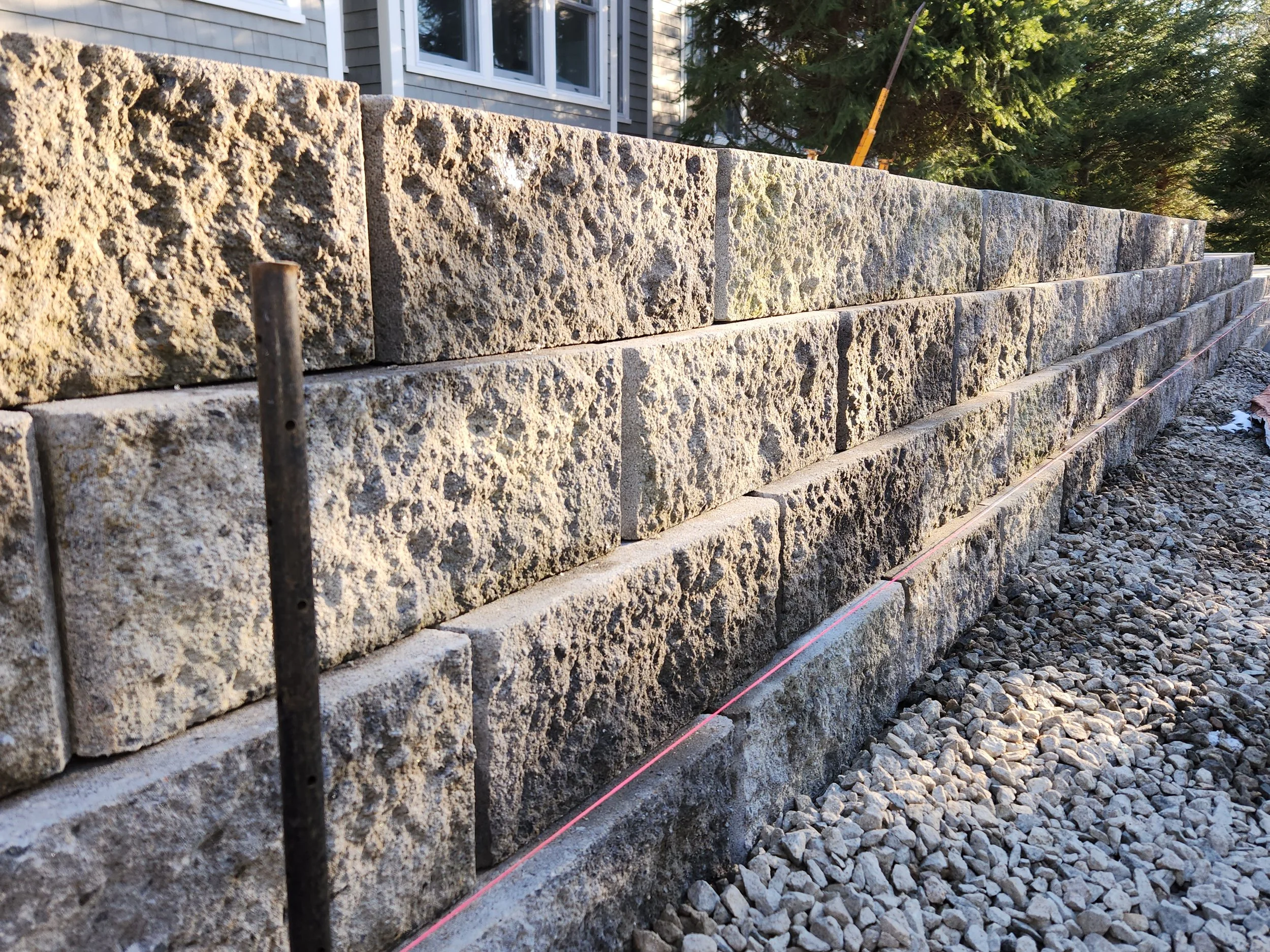 A stone retaining wall under construction with large, rough-cut stone blocks, a measuring string, and gravel at the base, with a house and trees in the background.