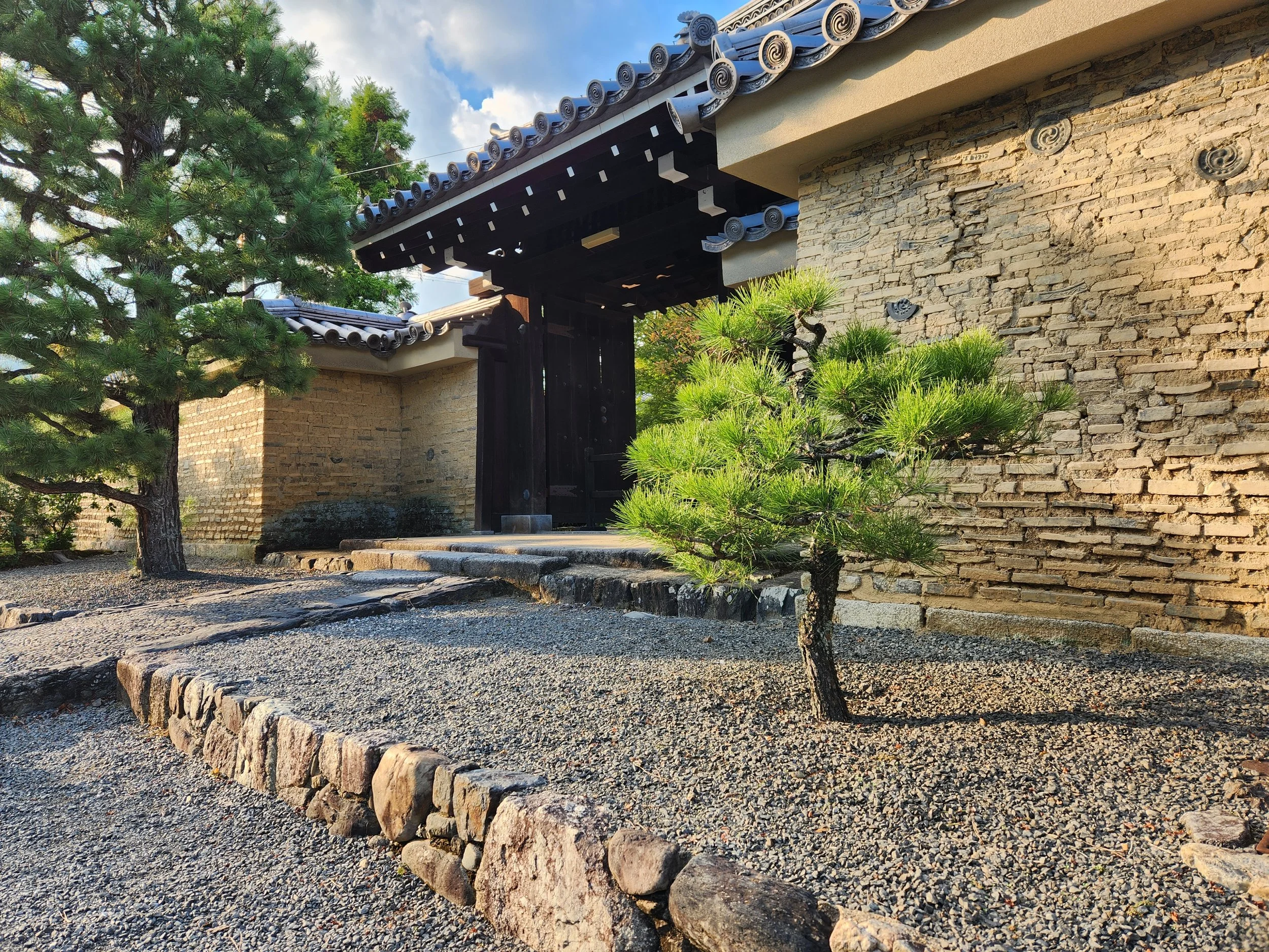 Traditional Japanese garden with a stone path, small pine trees, a brick wall, and a wooden gate under a tiled roof.