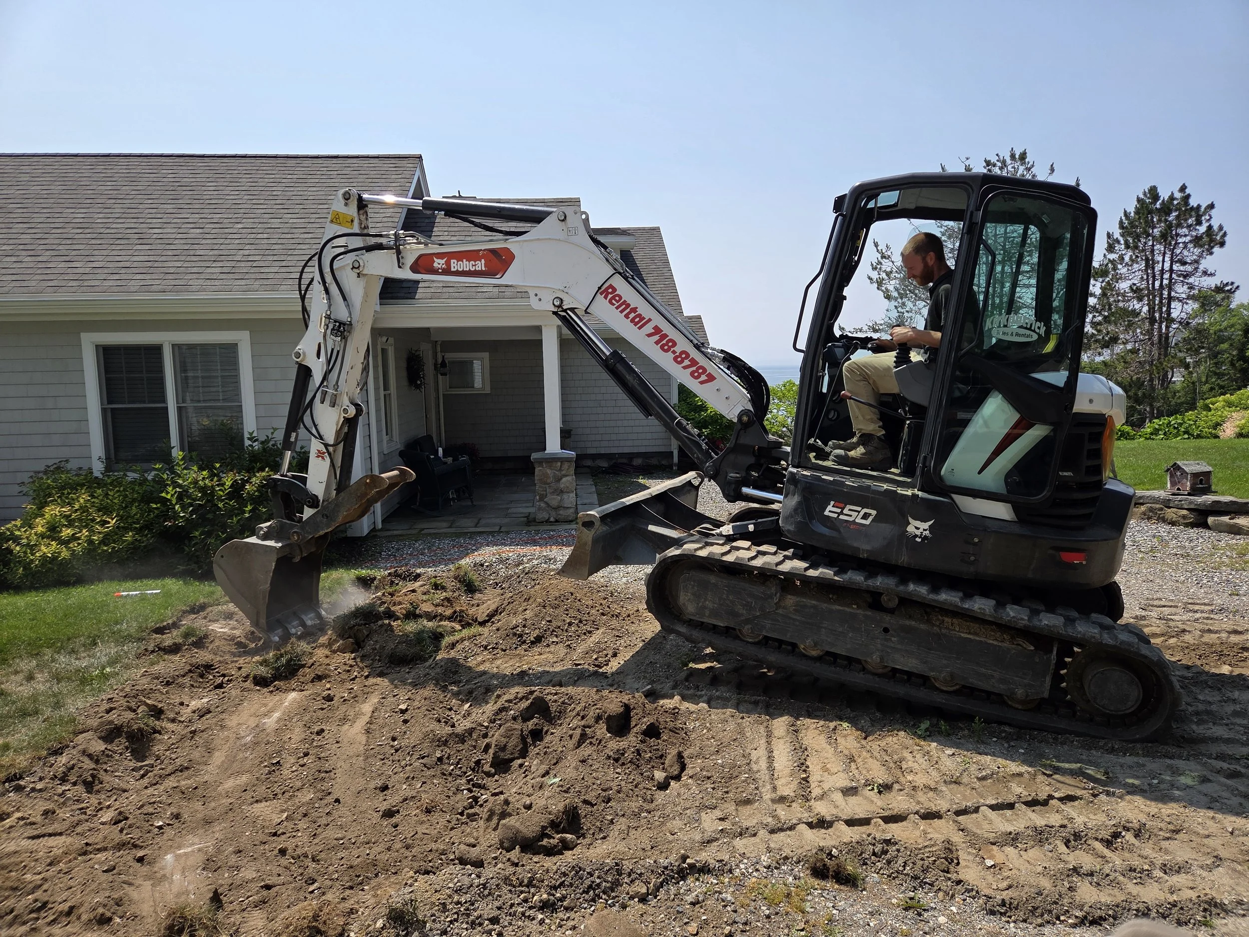 A man operating a small excavator digging a trench in a residential yard with a house and green lawn in the background.