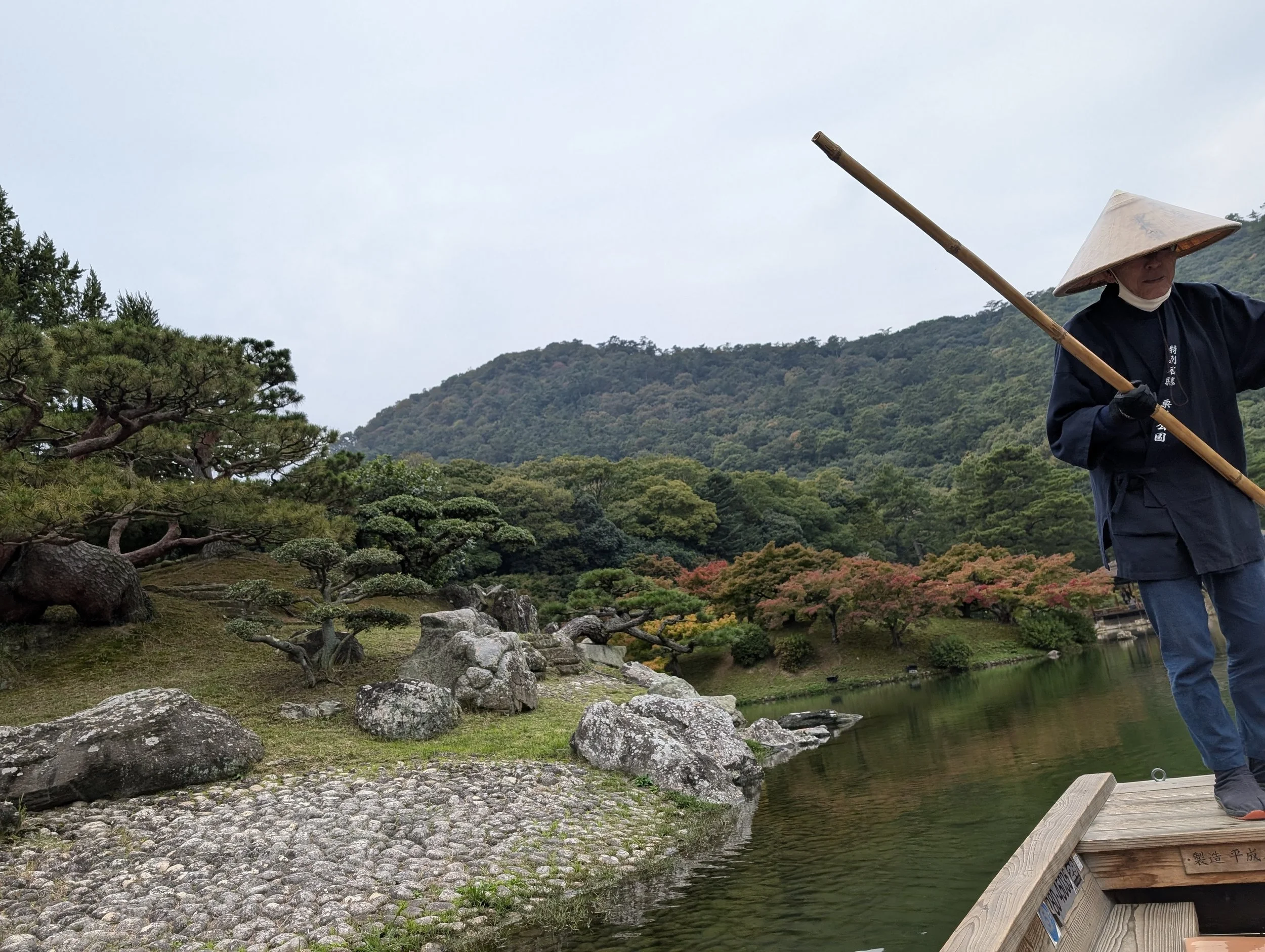 A person wearing traditional clothing and a conical hat, standing on a wooden boat with a pole, on a pond surrounded by rocks, trees, and mountains.