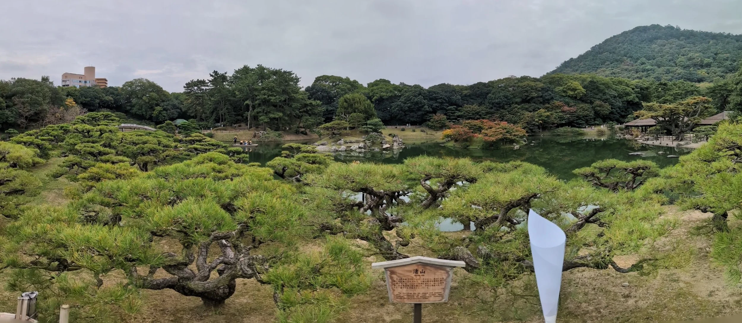 A scenic Japanese garden with trimmed pine trees, a pond, and distant structures amid lush greenery, with a mountain on the right and cloudy sky.