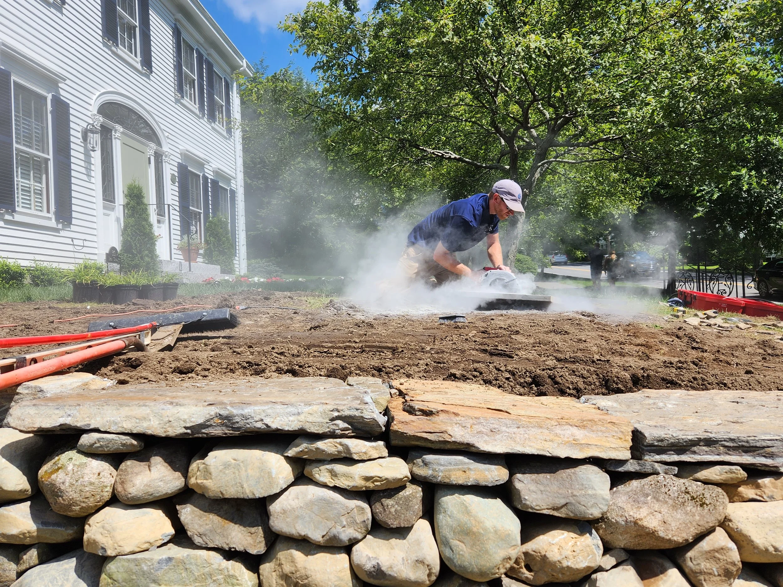 A man in a baseball cap and sunglasses uses a power saw to cut through a pathway under construction on a residential lawn, with dust and smoke in the air, and a stone wall in the foreground, a white house with black shutters in the background, and trees and parked cars around.