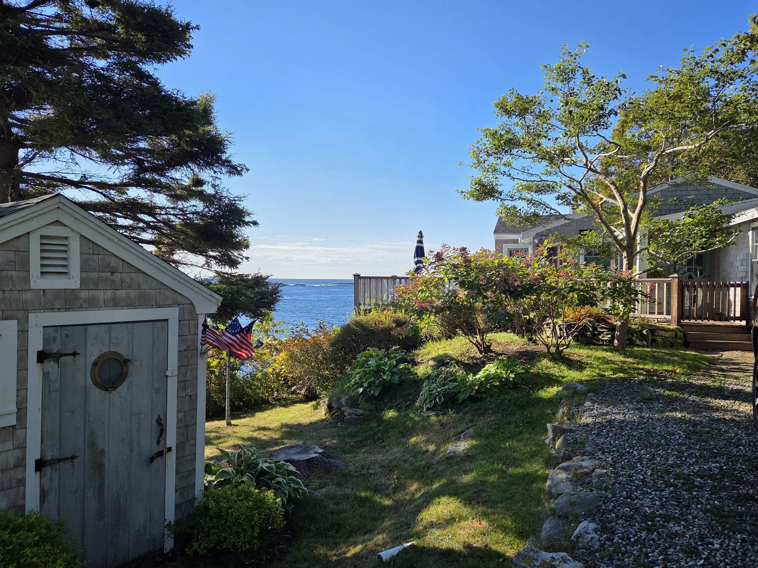 A backyard garden overlooking a body of water under a clear blue sky. There are trees, bushes, and flowering plants, a small shed with American flags, and a house with a deck.