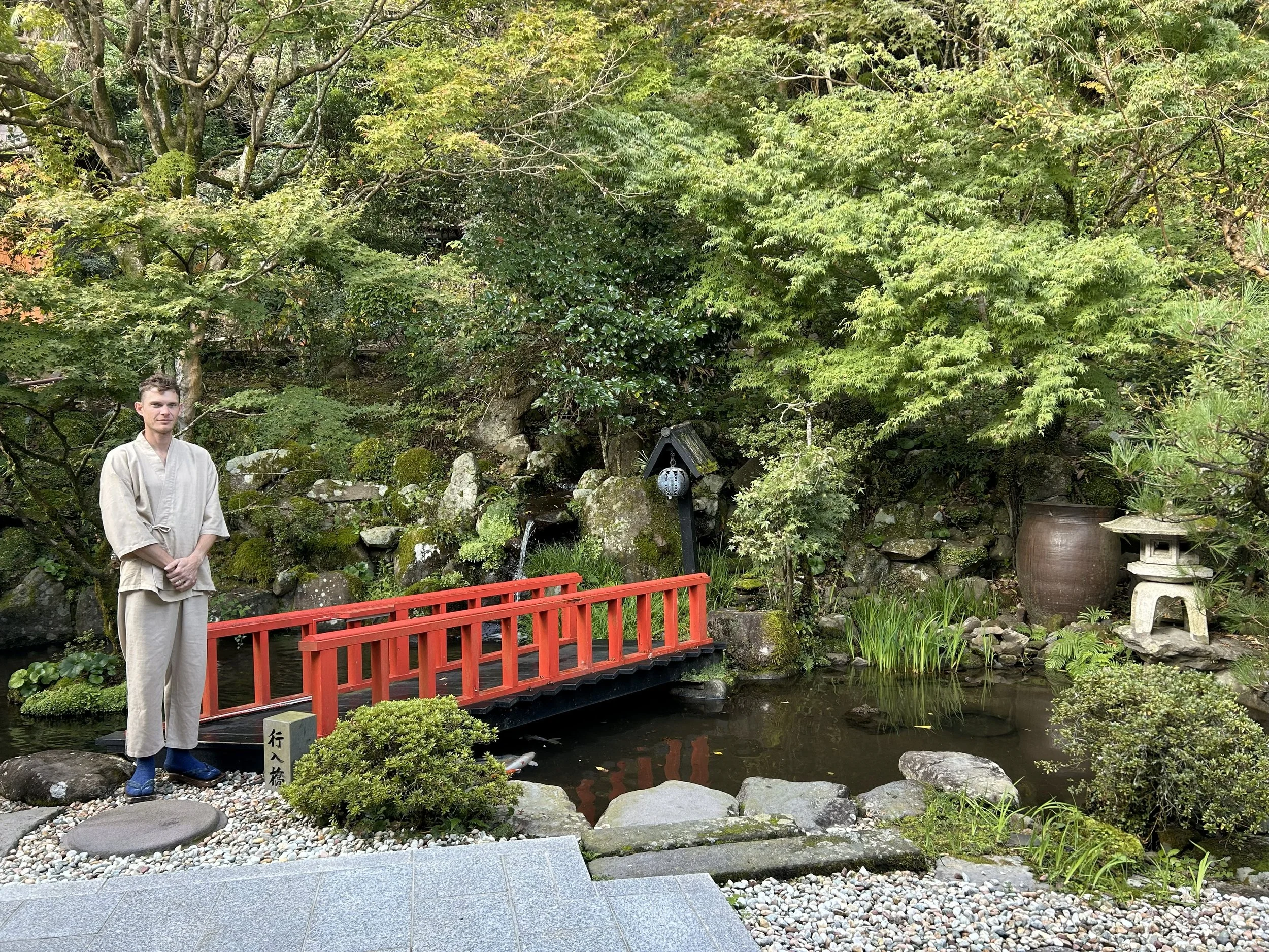 Person in traditional Japanese attire standing near a small pond in a Japanese garden with a red bridge, lush greenery, and ornamental objects.