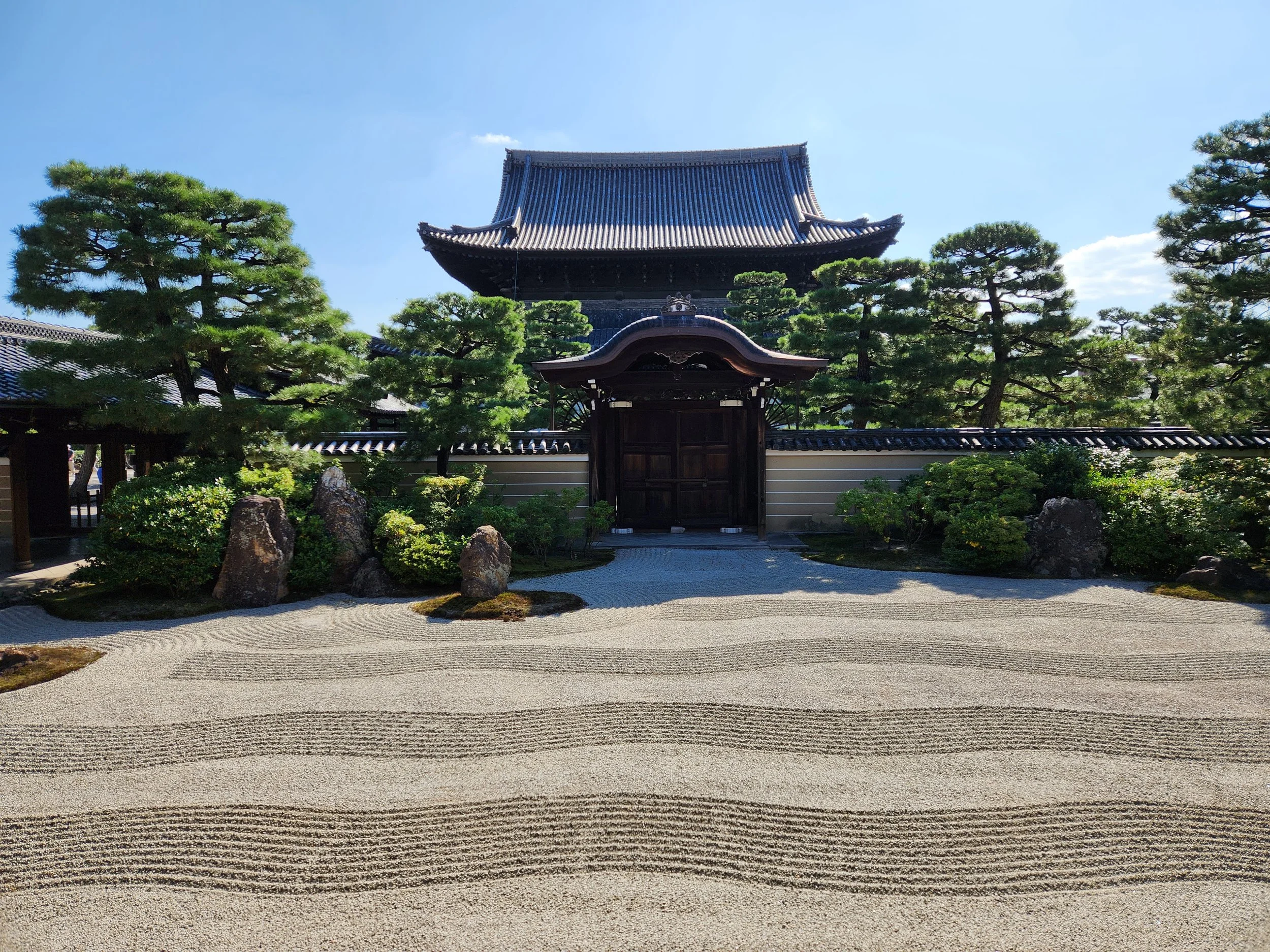 Traditional Japanese Zen garden with neatly raked gravel, large rocks, trimmed bushes, pine trees, and a wooden gate in front of a historic wooden building under a blue sky.