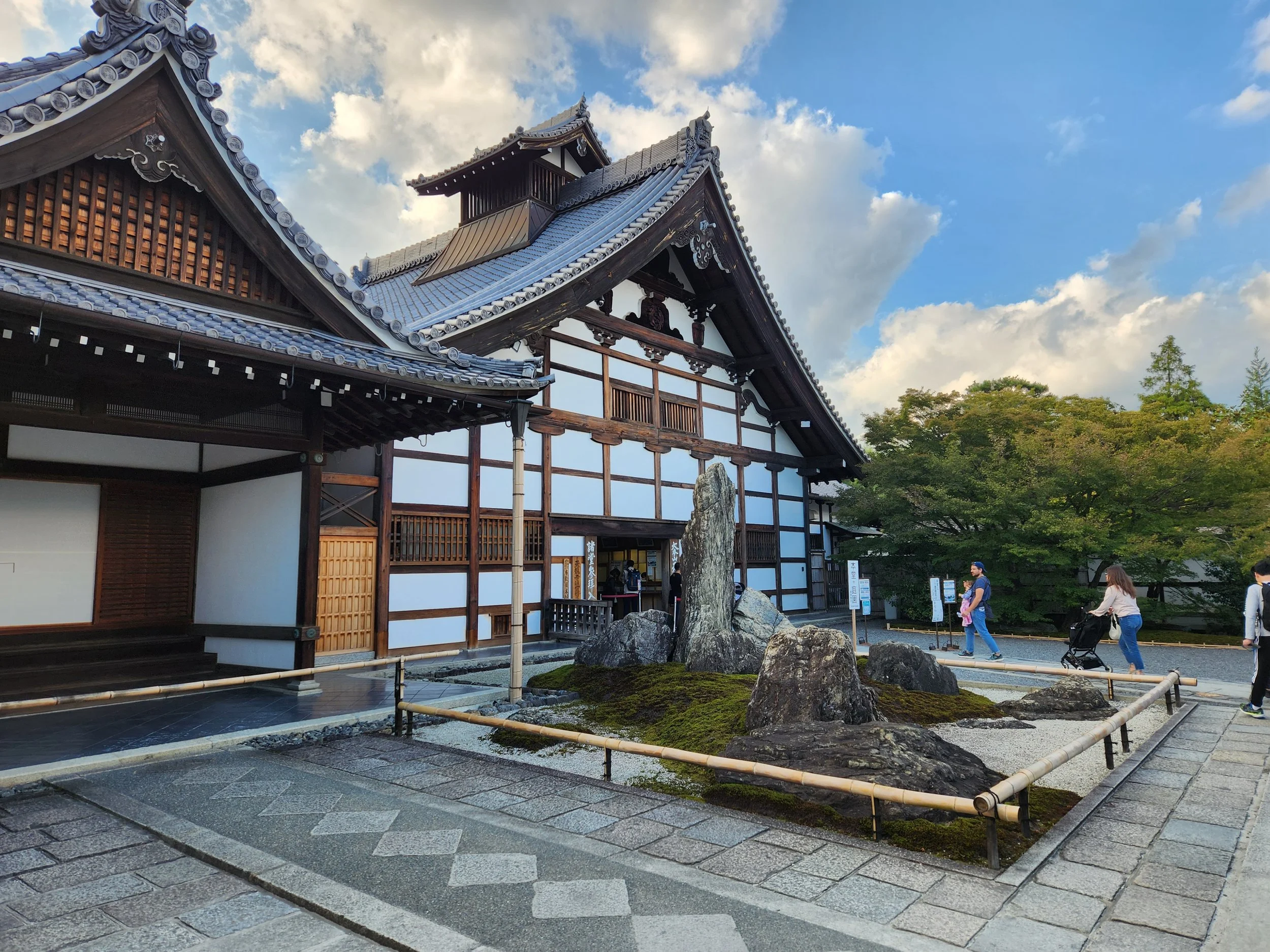 Traditional Japanese building with a rock garden in front, surrounded by a paved walkway and visitors walking nearby, under a partly cloudy sky.